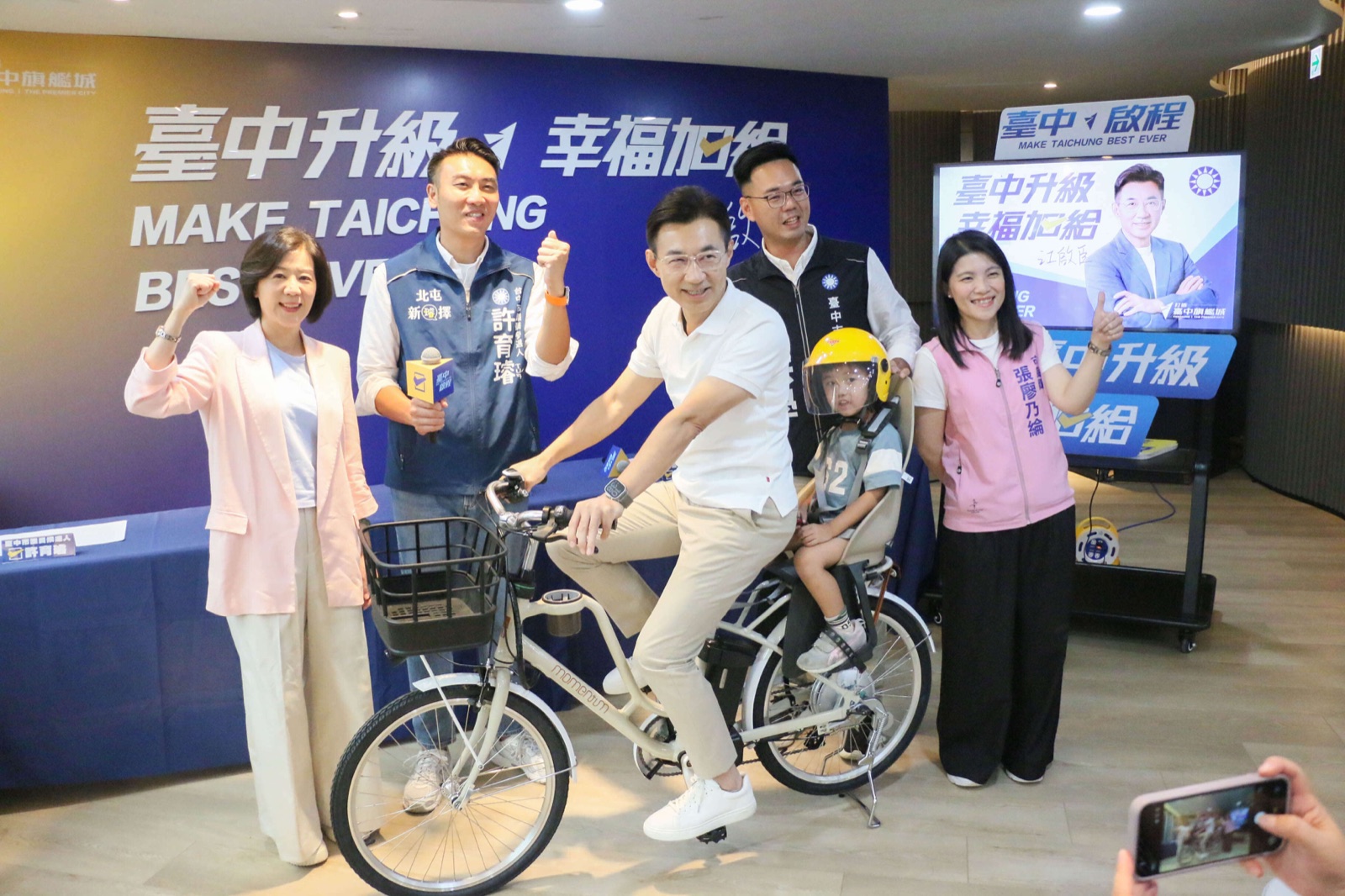 江啟臣在記者會現場試騎配備兒童安全座椅的「袋鼠車」共享單車，後座載著孩童