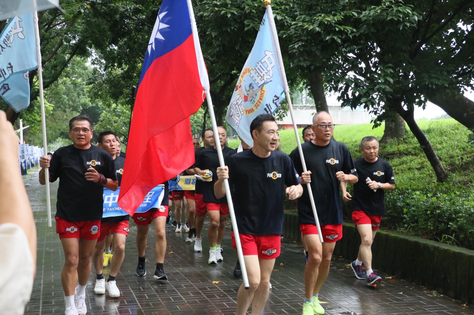 江啟臣手持中華民國國旗，與海龍退役弟兄在雨中沿人行道跑向登記會場