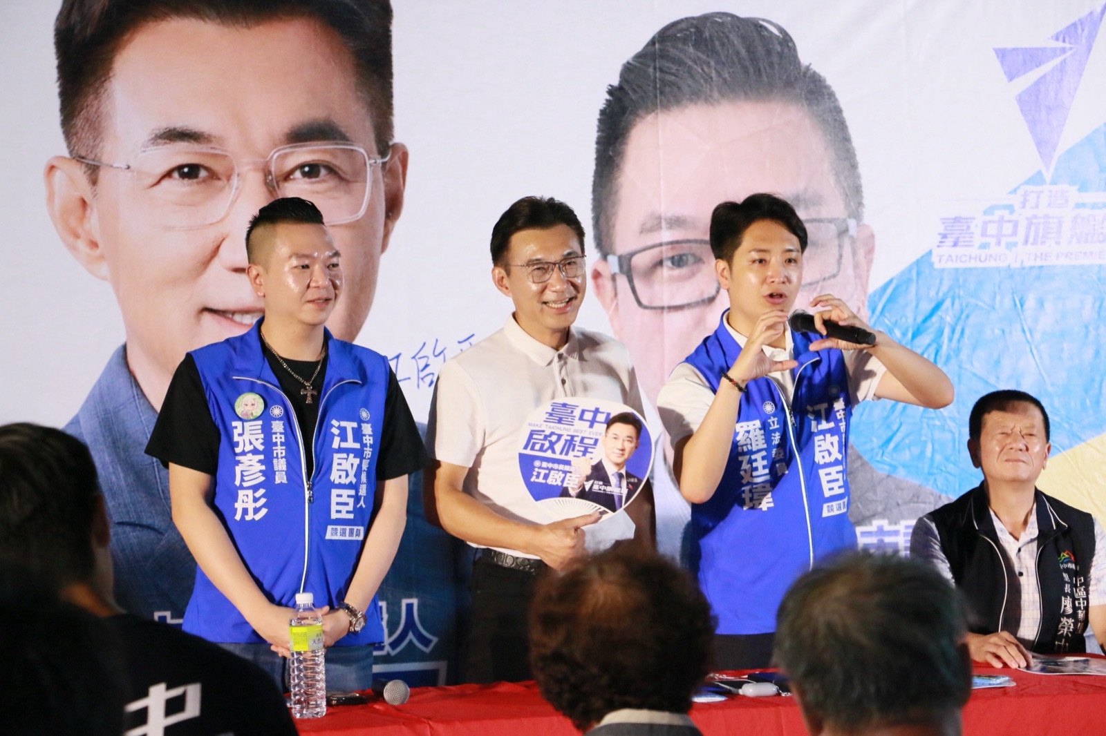羅廷瑋在台上以手比愛心發言，江啟臣手持「臺中啟程」扇子微笑，張彥彤在旁