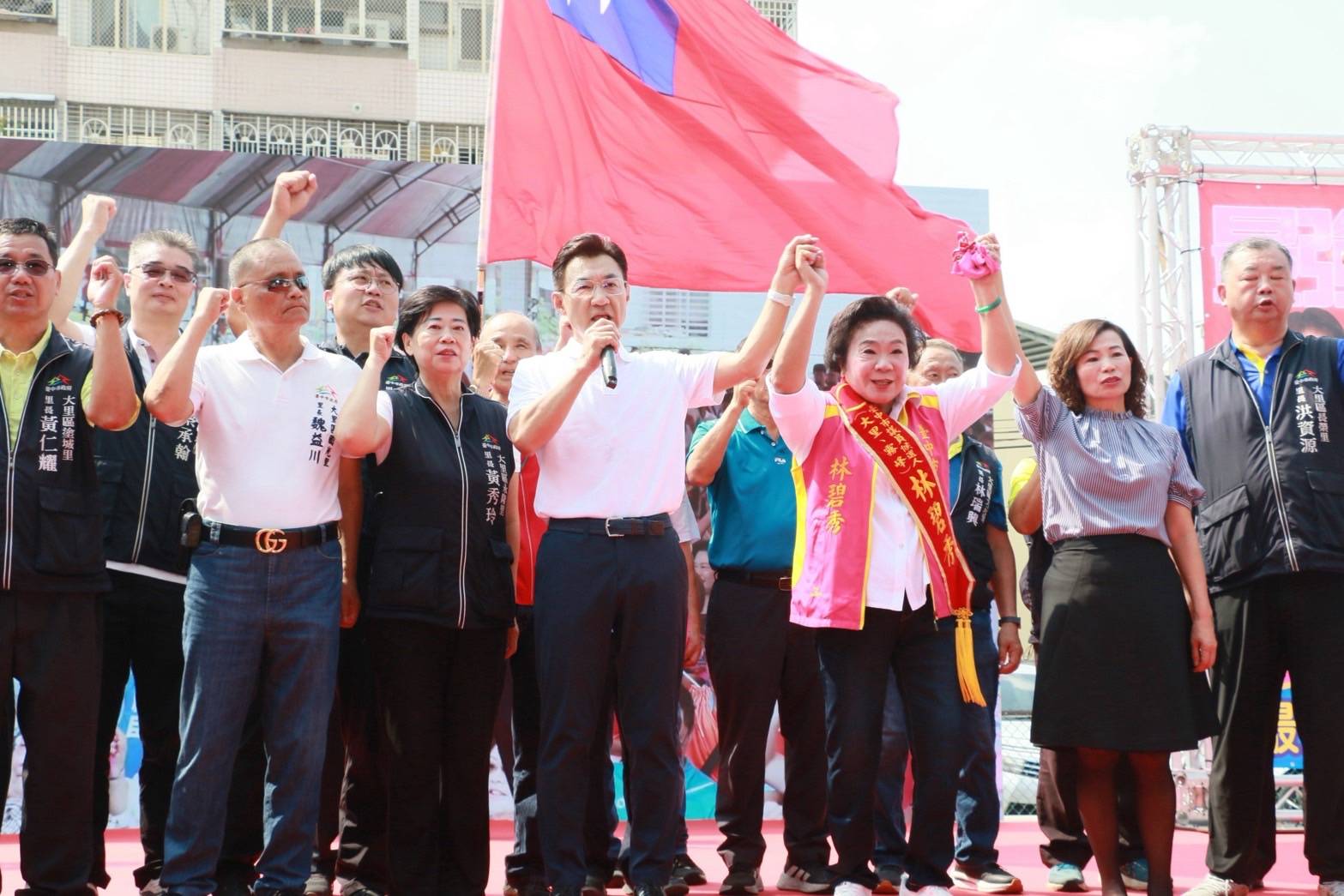 大里聯合競選總部成立大會舞台上，江啟臣持麥克風並牽起市議員林碧秀的手高舉，背景為中華民國國旗