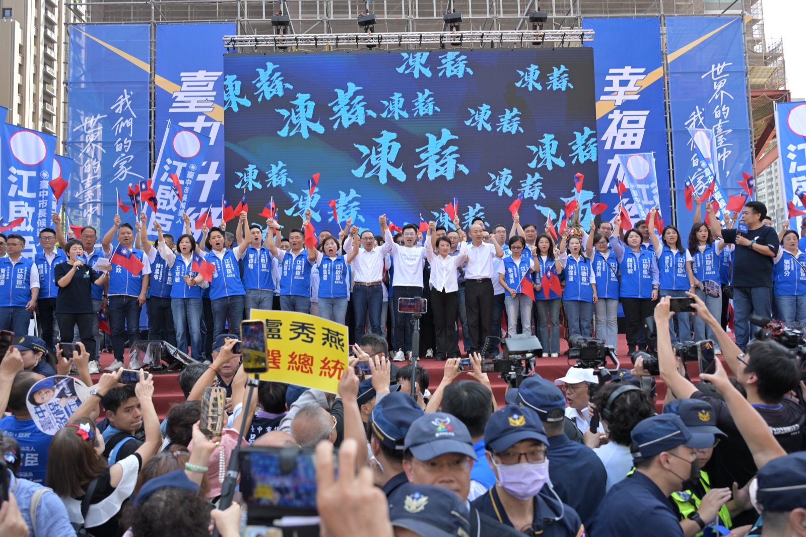 競選總部成立大會舞台，江啟臣與來賓手牽手高舉，台上立委與議員穿著藍色背心揮舞國旗，後方大螢幕滿是「凍蒜」