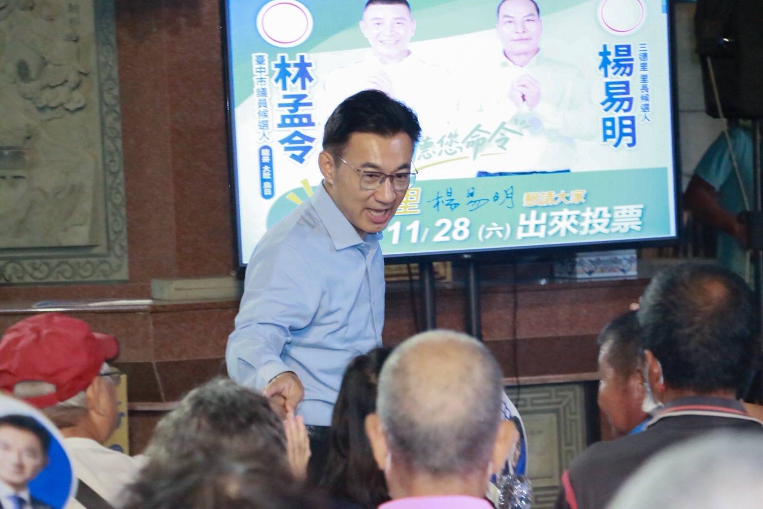 江啟臣在聖德宮與長輩握手，背後螢幕是林孟令與三德里長候選人楊易明的競選看板
