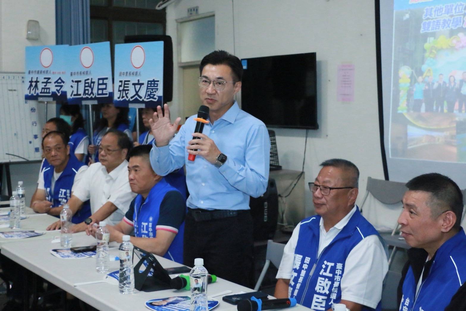 江啟臣在田中里活動中心手持麥克風說明政策，身後舉著林孟令、江啟臣、張文慶的候選人牌