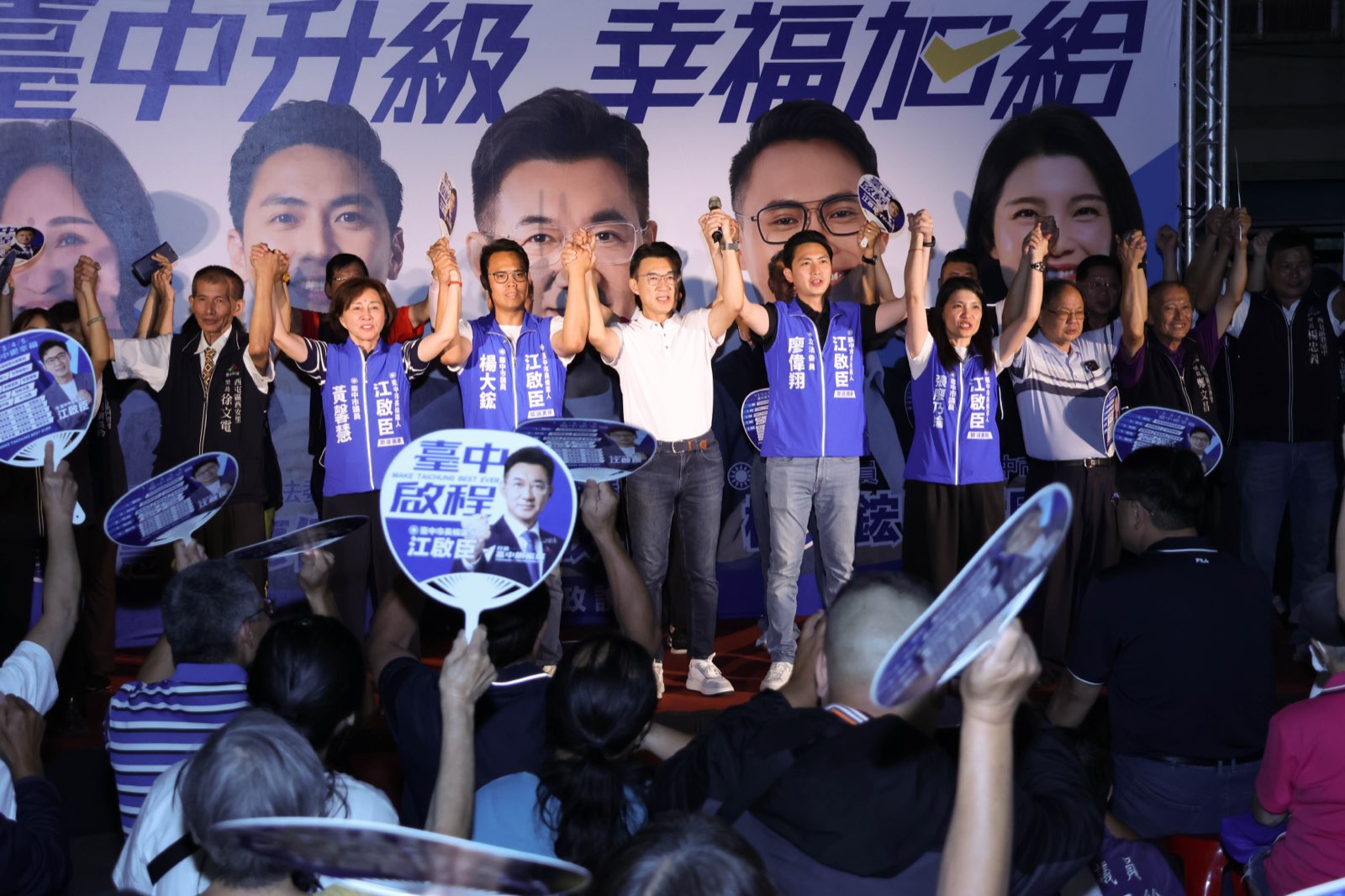 西屯問政說明會舞台上，江啟臣與廖偉翔、楊大鋐、張廖乃綸、黃馨慧及里長們牽手高舉，台下鄉親揮舞應援扇