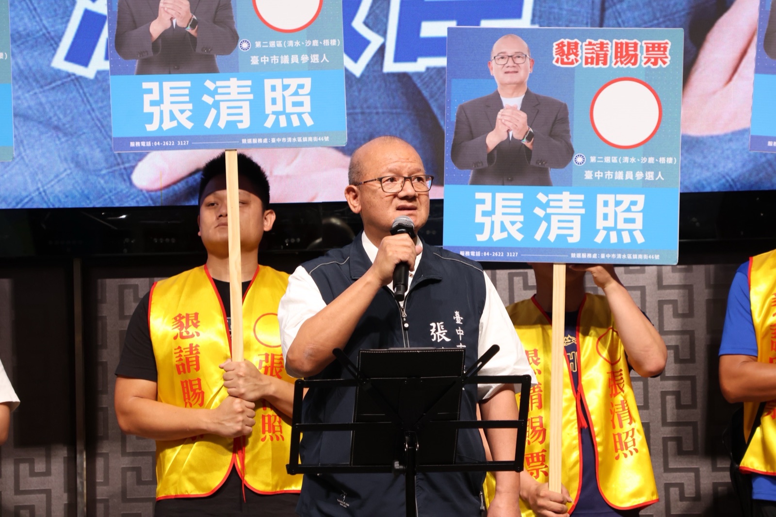穿深藍背心的男子手持麥克風發言，身後有人舉著第二選區（清水、沙鹿、梧棲）市議員參選人「張清照」看板，工作人員穿著「懇請賜票 張清照」黃背心