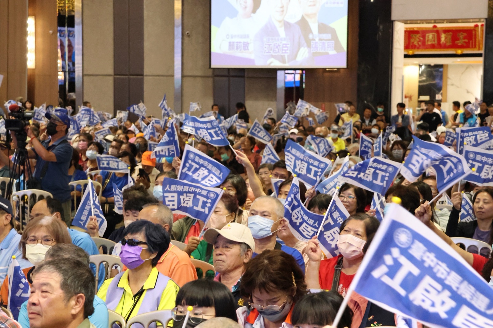 說明會現場座無虛席，鄉親揮舞寫著「臺中市長候選人 江啟臣」的藍色小旗，後方螢幕顯示顏莉敏、江啟臣、張清照三人