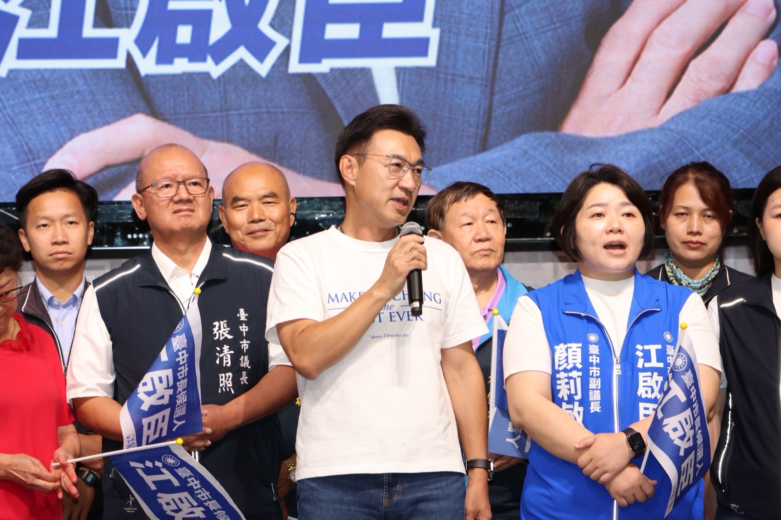 穿白T的男子手持麥克風發言，左側背心寫著「臺中市議長 張清照」，右側背心寫著「臺中市副議長 顏莉敏」，兩人手握江啟臣競選小旗