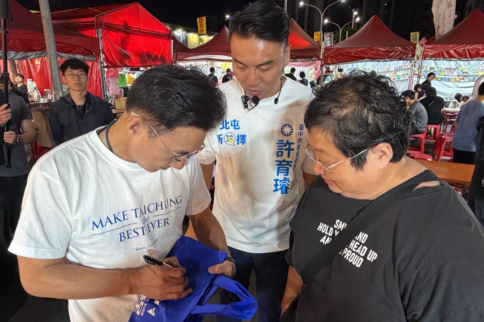 穿白T的男子在藍色提袋上簽名，一旁穿著「北屯 新擇」白T的人與一位民眾低頭觀看