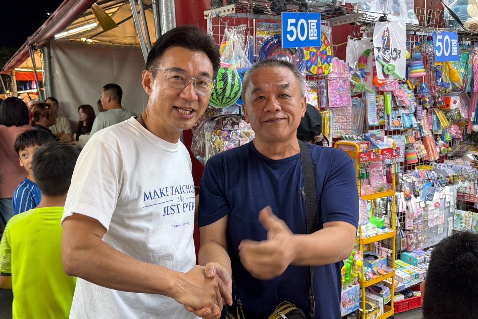 穿白T的男子在玩具攤前與攤商握手合照，攤位掛滿各式玩具