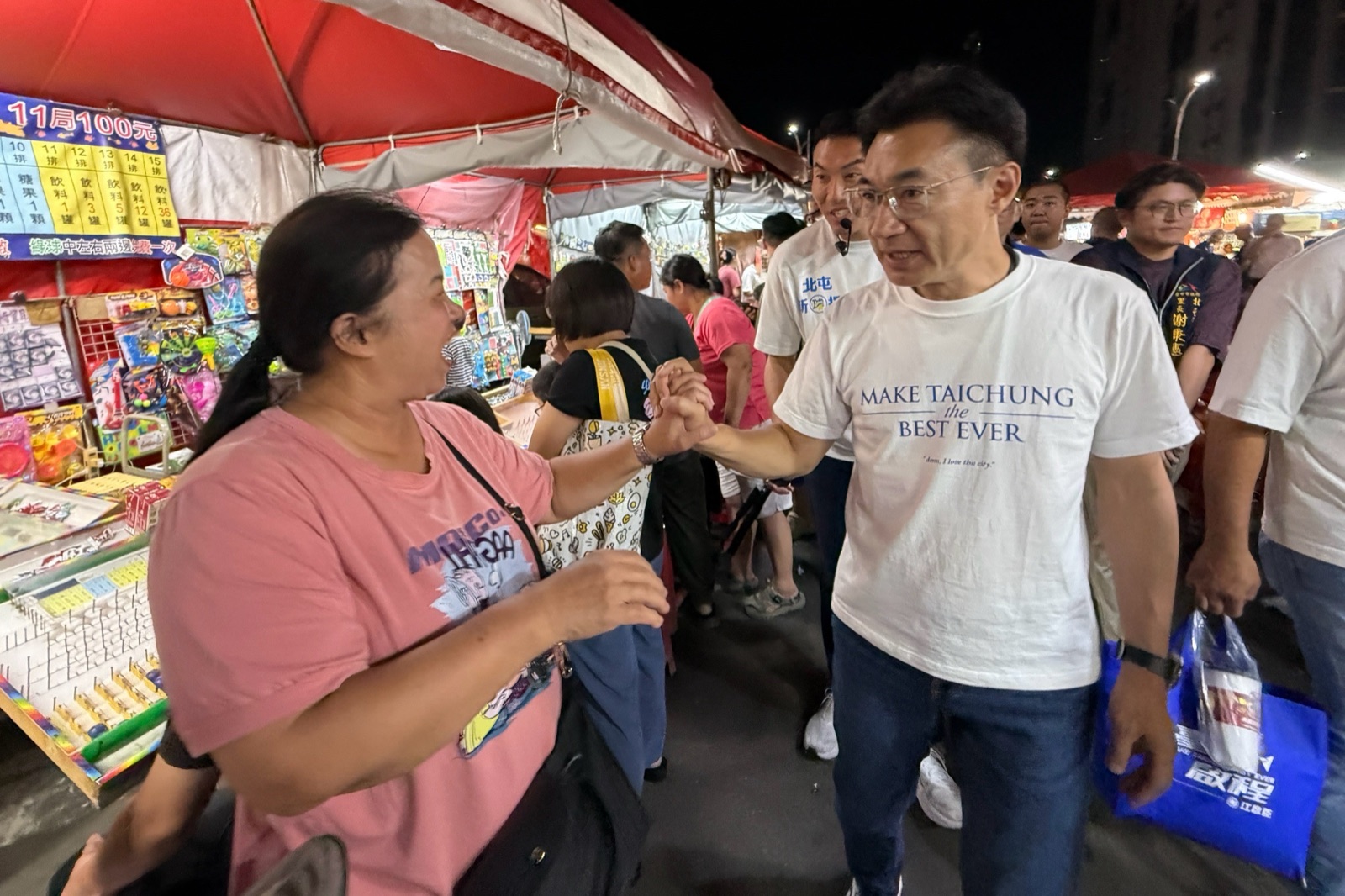 穿白T、提著藍色提袋的男子在夜市攤位間與一位攤商女士握手寒暄，身後有人陪同