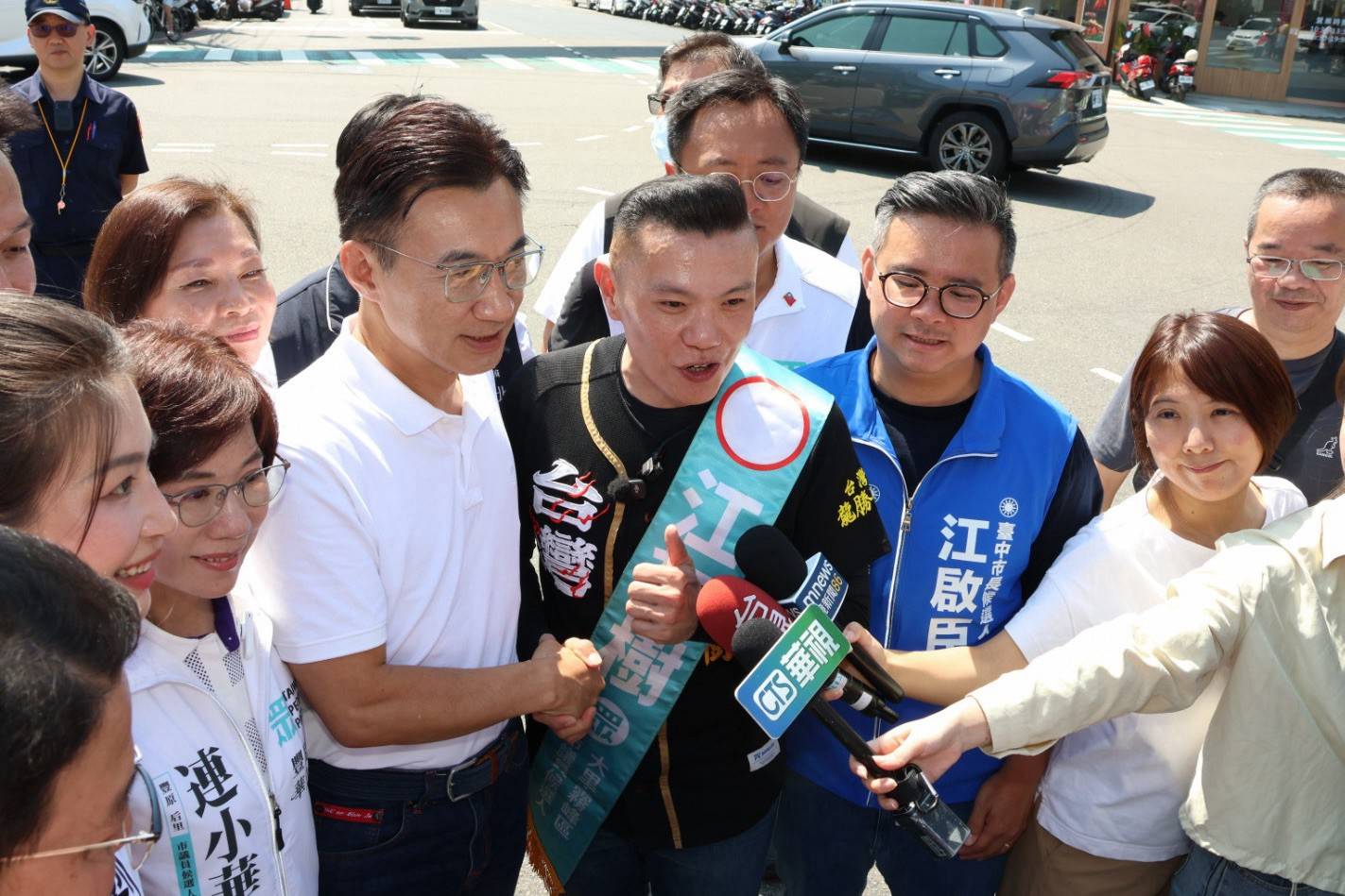穿白色Polo衫的男子與披青綠色候選人肩帶、比讚的男子握手，記者伸出多支麥克風採訪，周圍有穿「江啟臣 競選團隊」藍背心的人