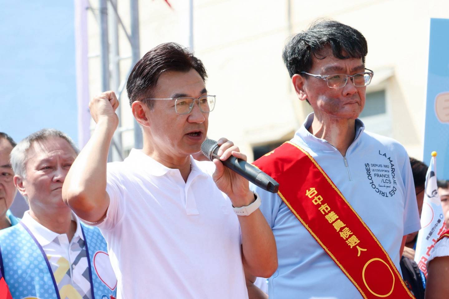 穿白色Polo衫的男子握拳、手持麥克風發言，旁邊穿淺藍Polo衫、披「市議員候選人」肩帶的男子在一旁聆聽