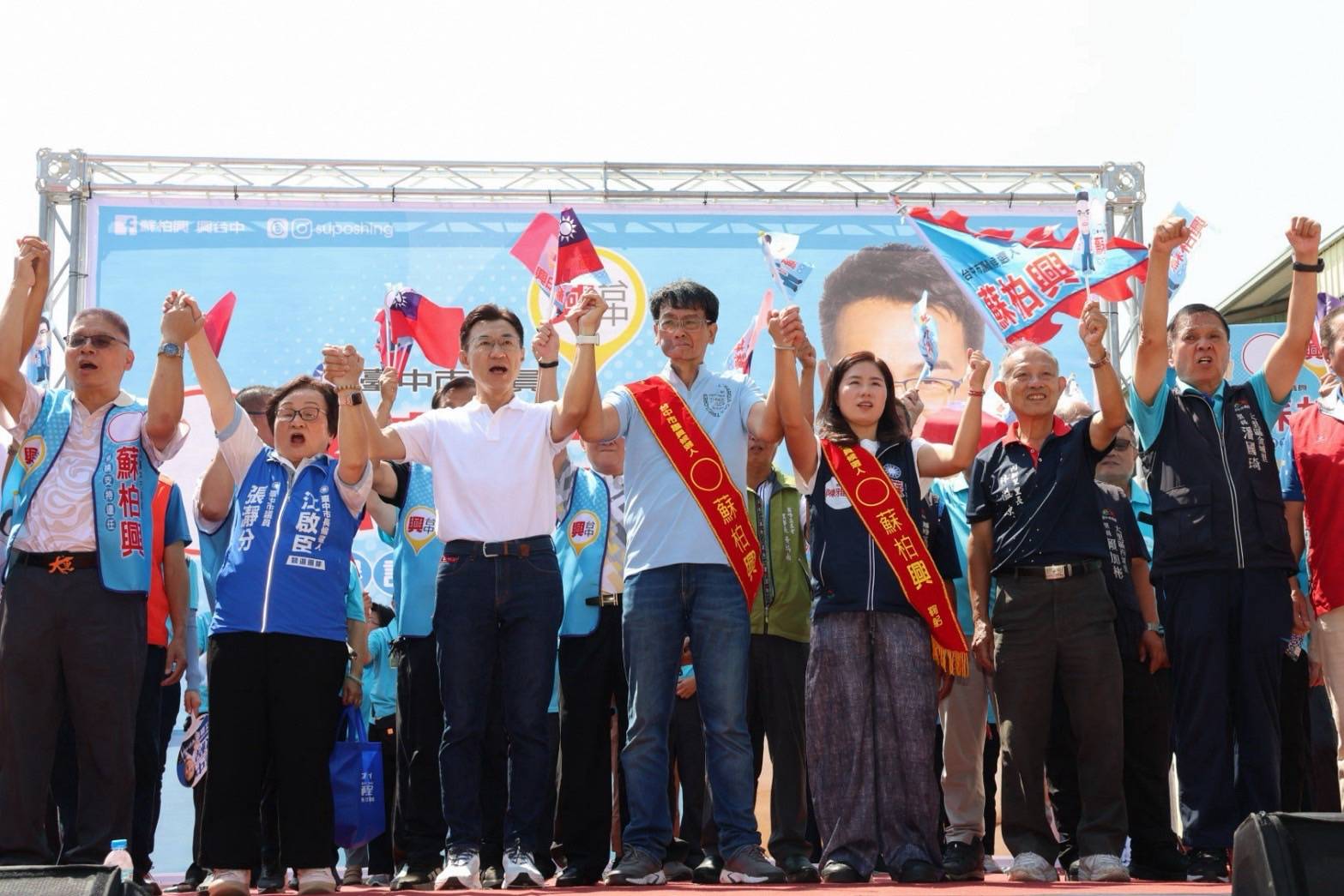 大里蘇柏興競選總部成立大會，臺上眾人手牽手高舉雙手，兩人披著寫有「蘇柏興」的肩帶，旗幟與背板上印著「蘇柏興」