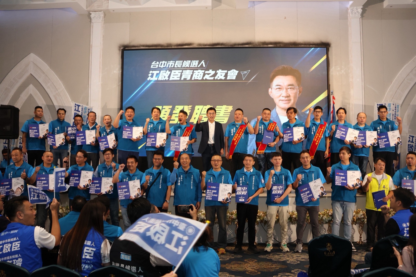 江啟臣青商之友會成立大會，穿青藍色背心的成員手持聘書、握拳站成兩排，居中穿深色西裝的男子與眾人合影，背後大螢幕寫著「江啟臣青商之友會」與「頒發聘書」