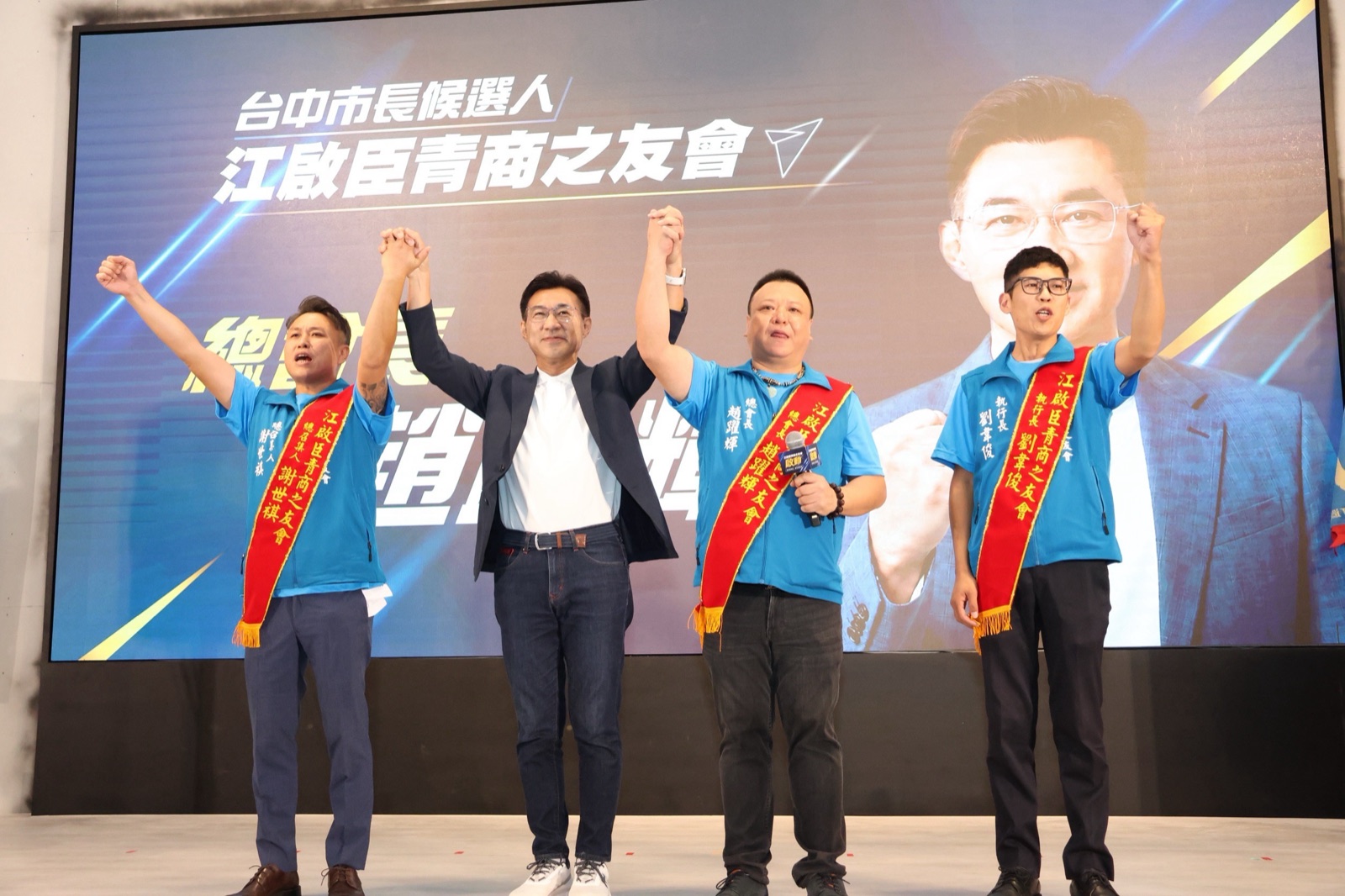 三位披「江啟臣青商之友會」紅色肩帶的幹部與一名穿深色外套的男子在臺上手牽手高舉，肩帶分別寫著「總召集人」「總會長」「執行長」