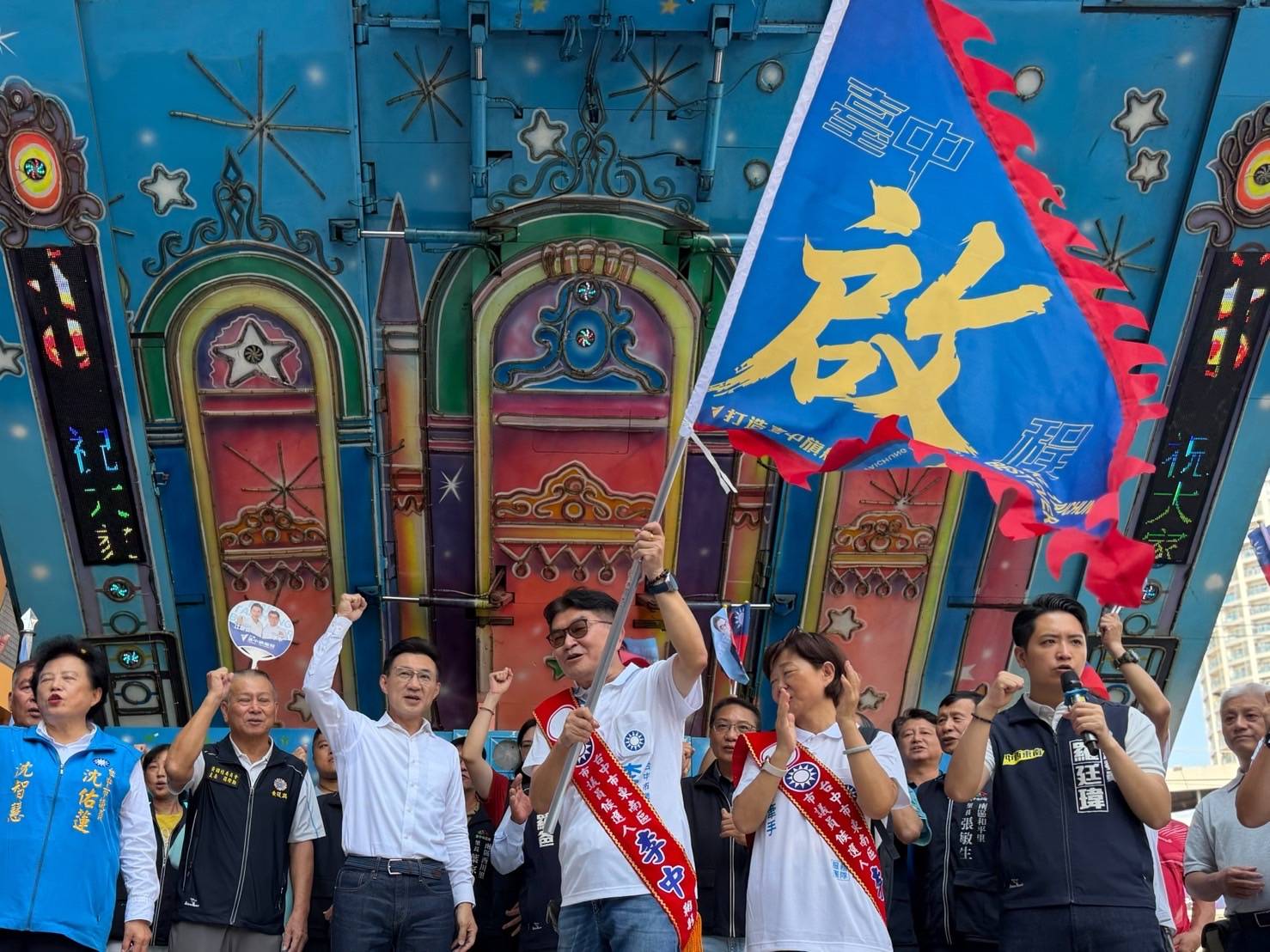 東南區競總成立現場，肩帶寫著「李中」的市議員候選人高舉揮舞藍底「臺中啟程」旗幟，臺上眾人握拳歡呼，右側穿「羅廷瑋」背心者手持麥克風