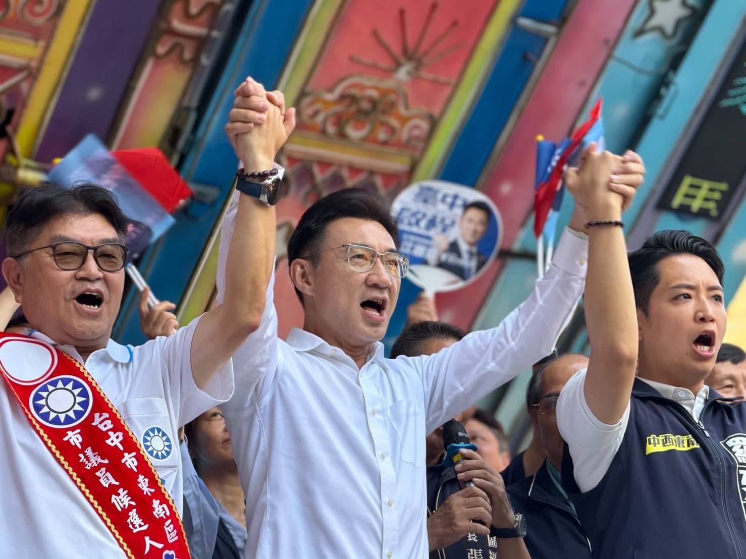 東南區競總成立現場，臺上三人牽手高舉雙手同聲高喊，左側披「東南區市議員候選人」肩帶，右側穿「中西東南」背心，背後有人揮舞國旗與競選扇