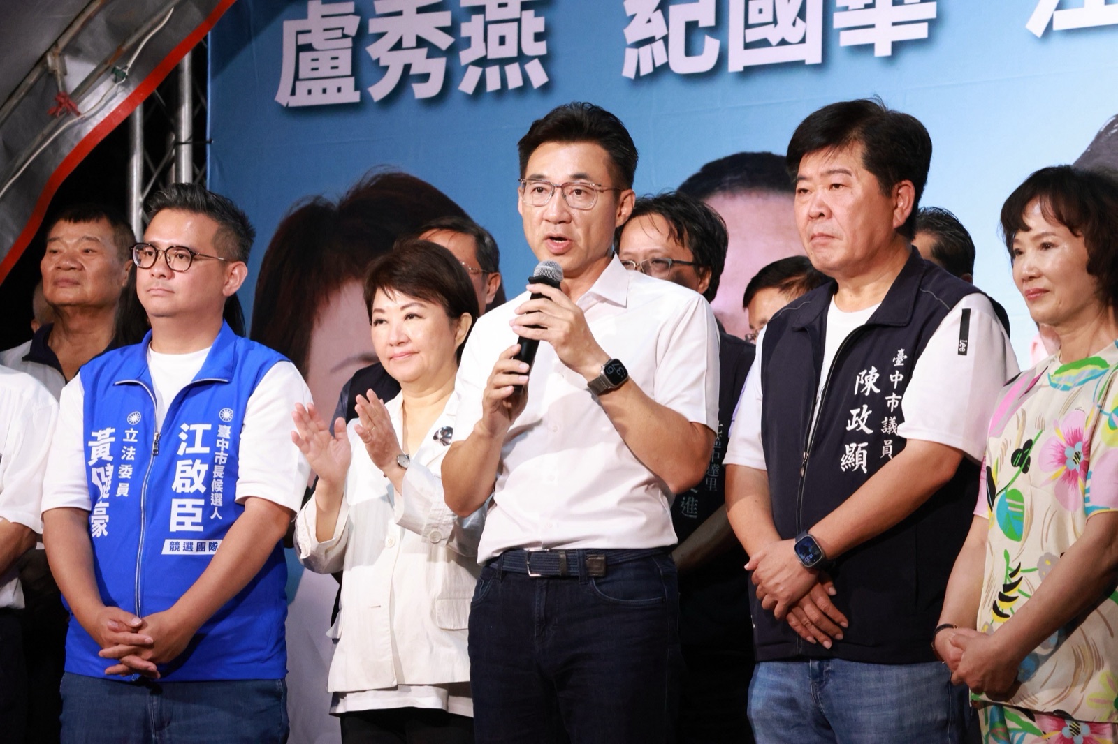 穿白襯衫的男子手持麥克風發言，左側背心寫著「立法委員 黃健豪」與「江啟臣 競選團隊」，右側背心寫著「臺中市議員 陳政顯」，一旁穿白外套的女士鼓掌
