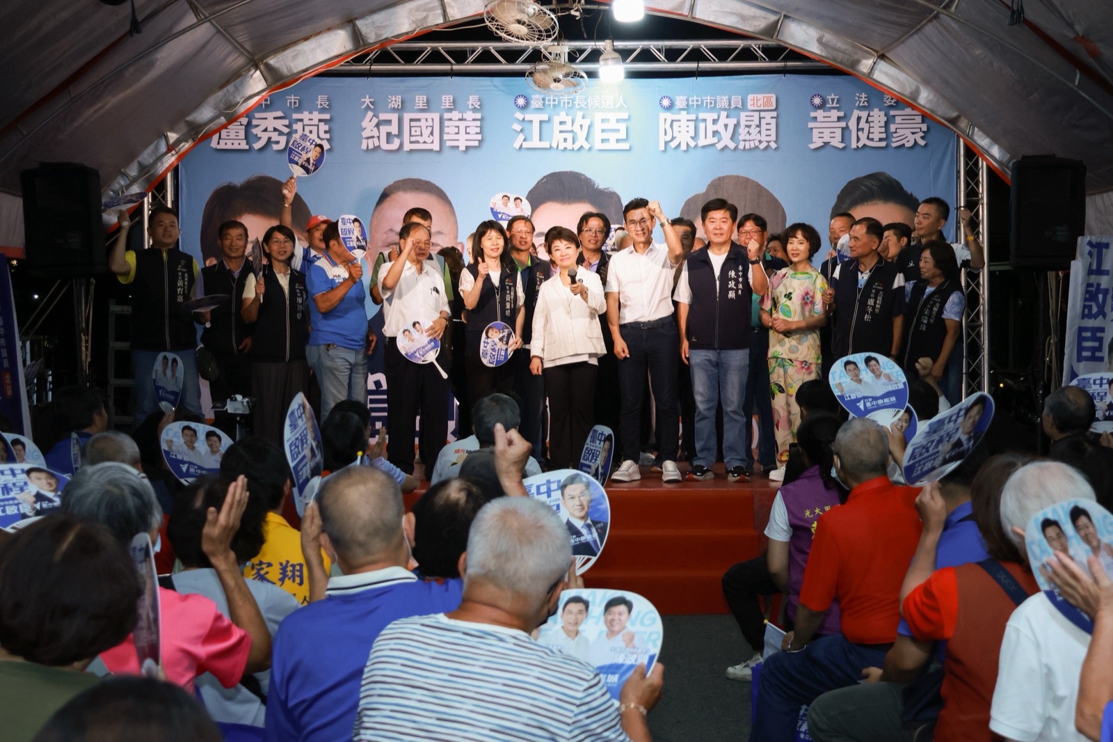 北區聯合問政說明會臺上眾人合影，背板列著「臺中市長 盧秀燕」「臺中市長候選人 江啟臣」「臺中市議員 陳政顯」「立法委員 黃健豪」等字樣，臺下鄉親高舉競選扇