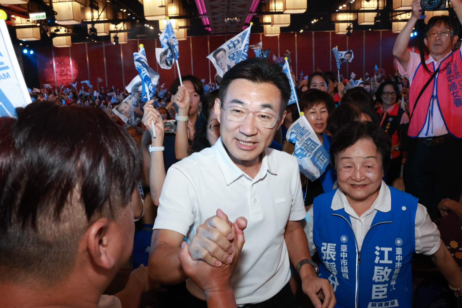 穿白色Polo衫的男子在走道上與民眾握手，身旁穿「臺中市議員 張靜分」背心的女士微笑，後方有人穿著「大臺中婦女會」背心