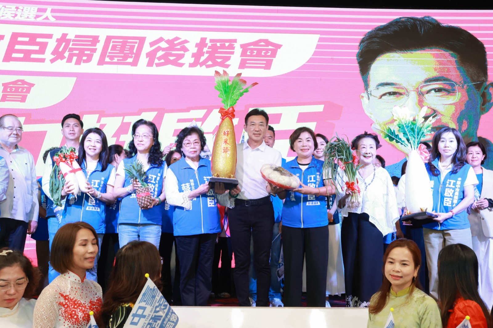 臺上婦團代表捧著寫有「好采頭」的大蘿蔔、鳳梨、青蔥與禮籃等吉祥物，與穿白襯衫的男子合影，背後螢幕寫著「婦團後援會」