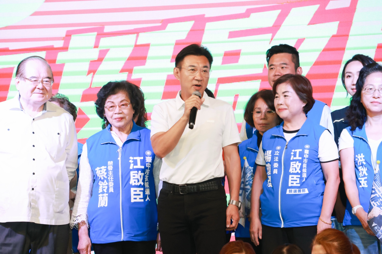 穿白色Polo衫的男子手持麥克風發言，左側背心寫著「榮譽主任委員 蔡鈴蘭」，右側背心寫著「立法委員 楊瓊瓔」