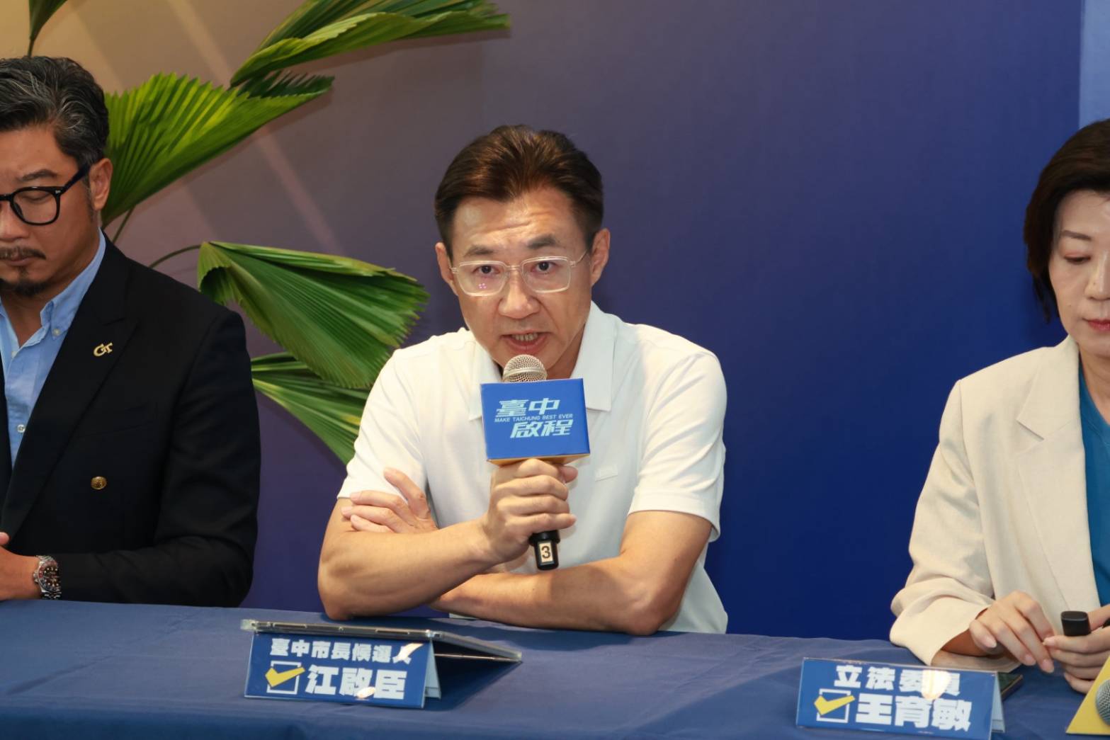 穿白色Polo衫的男子手持麥克風發言，桌上立牌寫著「臺中市長候選人 江啟臣」，右側桌牌寫著「立法委員 王育敏」