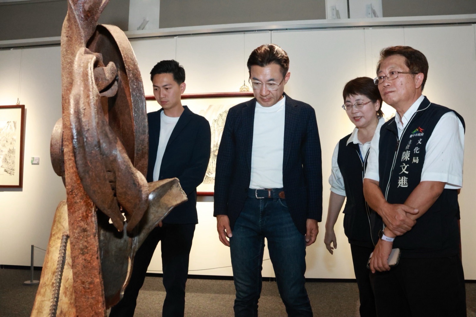 穿深藍西裝外套的男子與身旁人員在展間凝視一件鏽蝕的金屬雕塑，右側背心寫著「文化局 主任 陳文進」