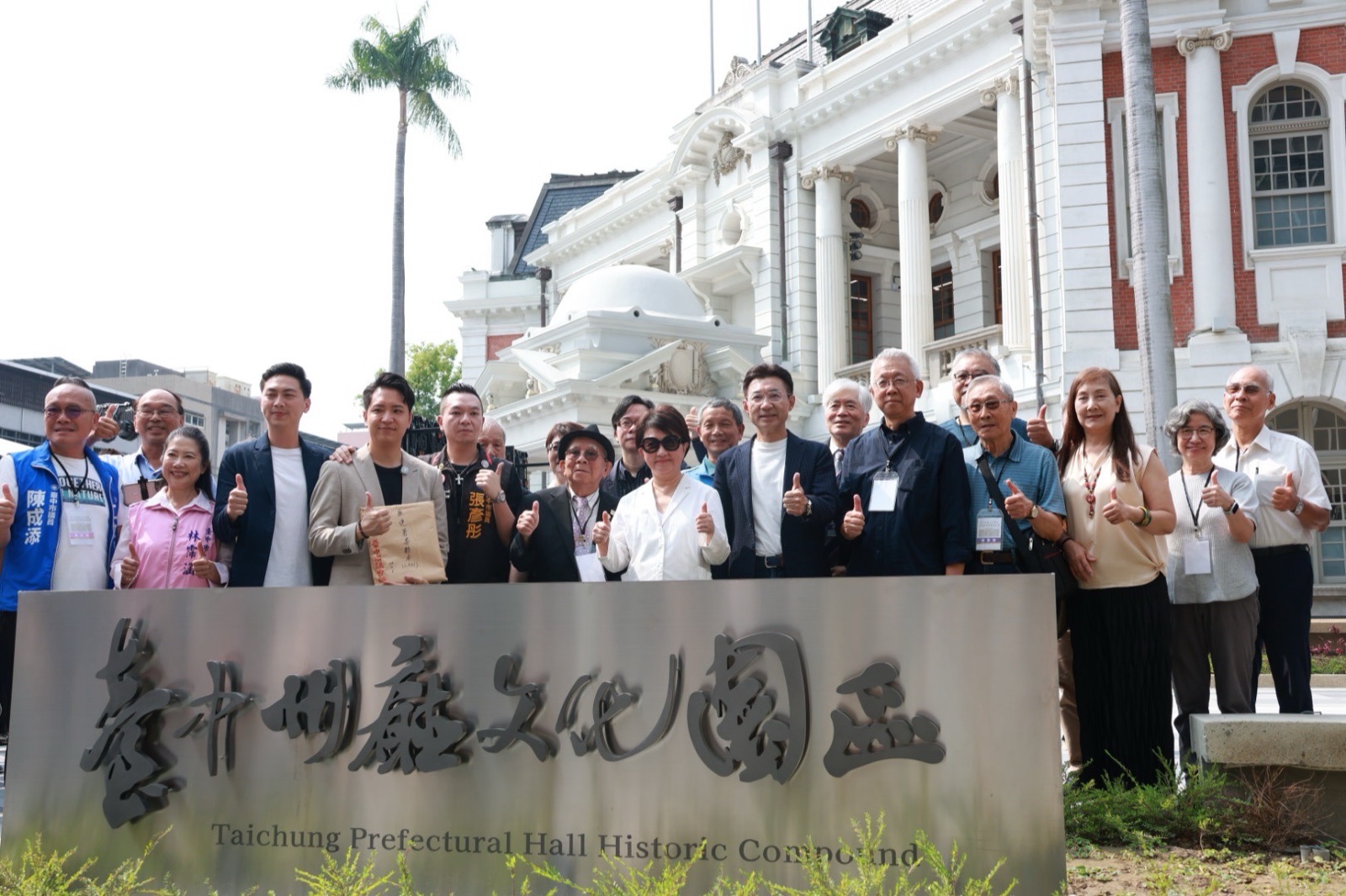 臺中州廳文化園區揭牌後的合影，眾人在「臺中州廳文化園區」不鏽鋼立牌後比讚，身後是白色古典風格的州廳主樓