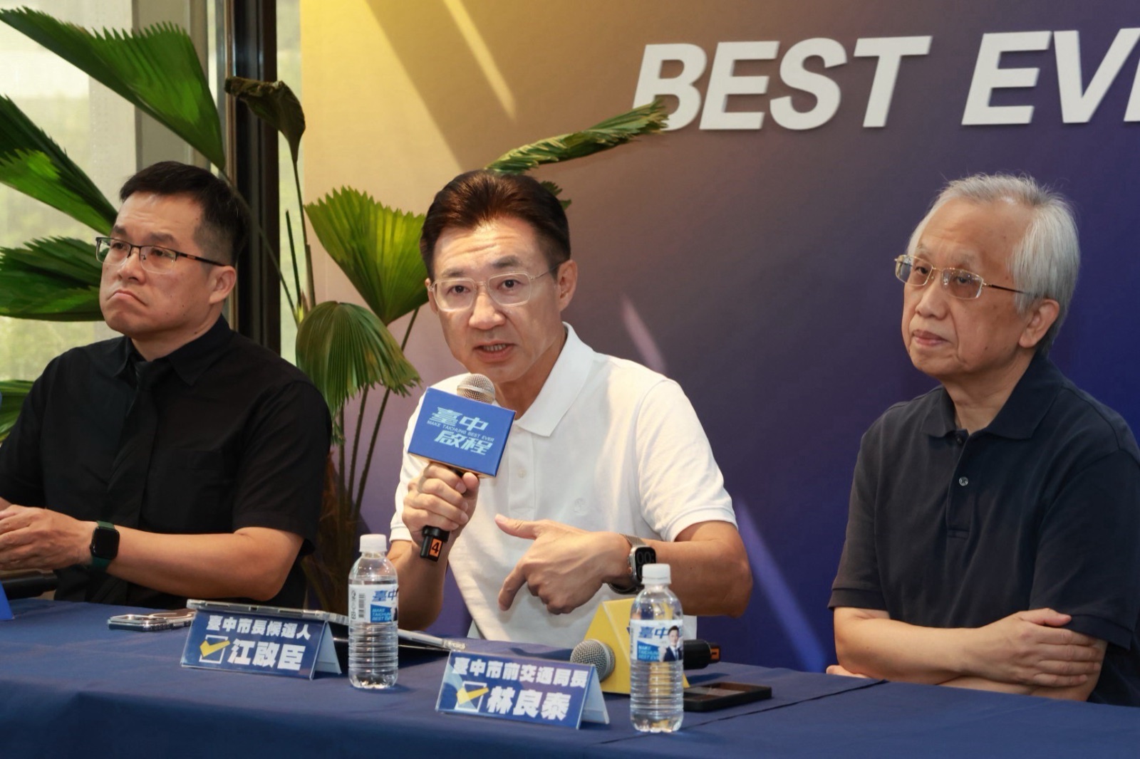 穿白色Polo衫的男子手持麥克風發言，桌上立牌寫著「臺中市長候選人 江啟臣」與「臺中市前交通局長 林良泰」
