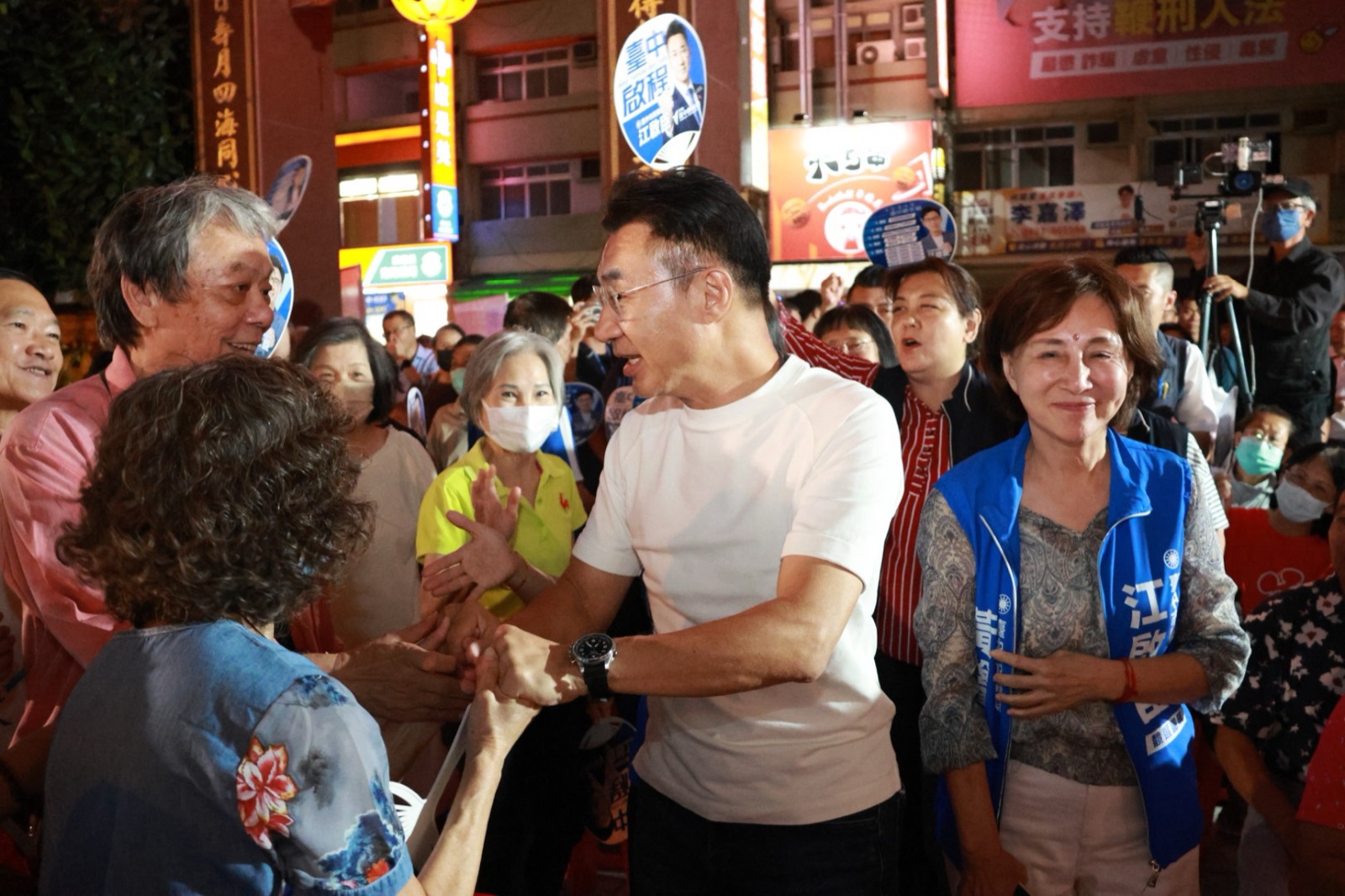 穿白T的男子與一位長輩握手，右側穿藍色「江啟臣」背心的女士微笑，後方鄉親舉著競選扇