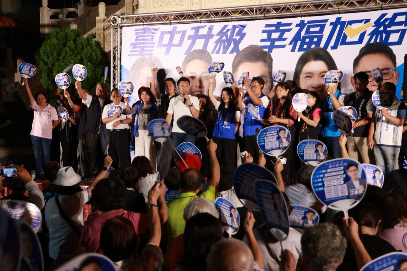 夜間廟前舞臺上，穿白T的男子手持麥克風，臺上民意代表與里長高舉競選扇，臺下鄉親舉扇回應，背板寫著「臺中升級 幸福加給」