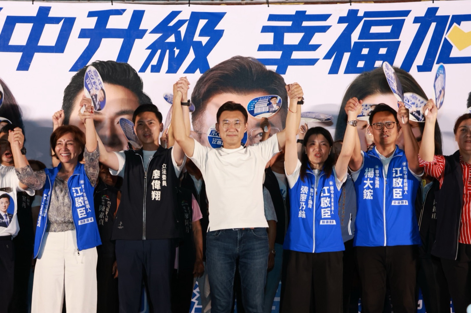 西屯問政說明會臺上眾人手牽手高舉雙手，背心寫著「立法委員 廖偉翔」「臺中市議員 張廖乃綸」「楊大鋐」，背後看板寫著「臺中升級 幸福加給」