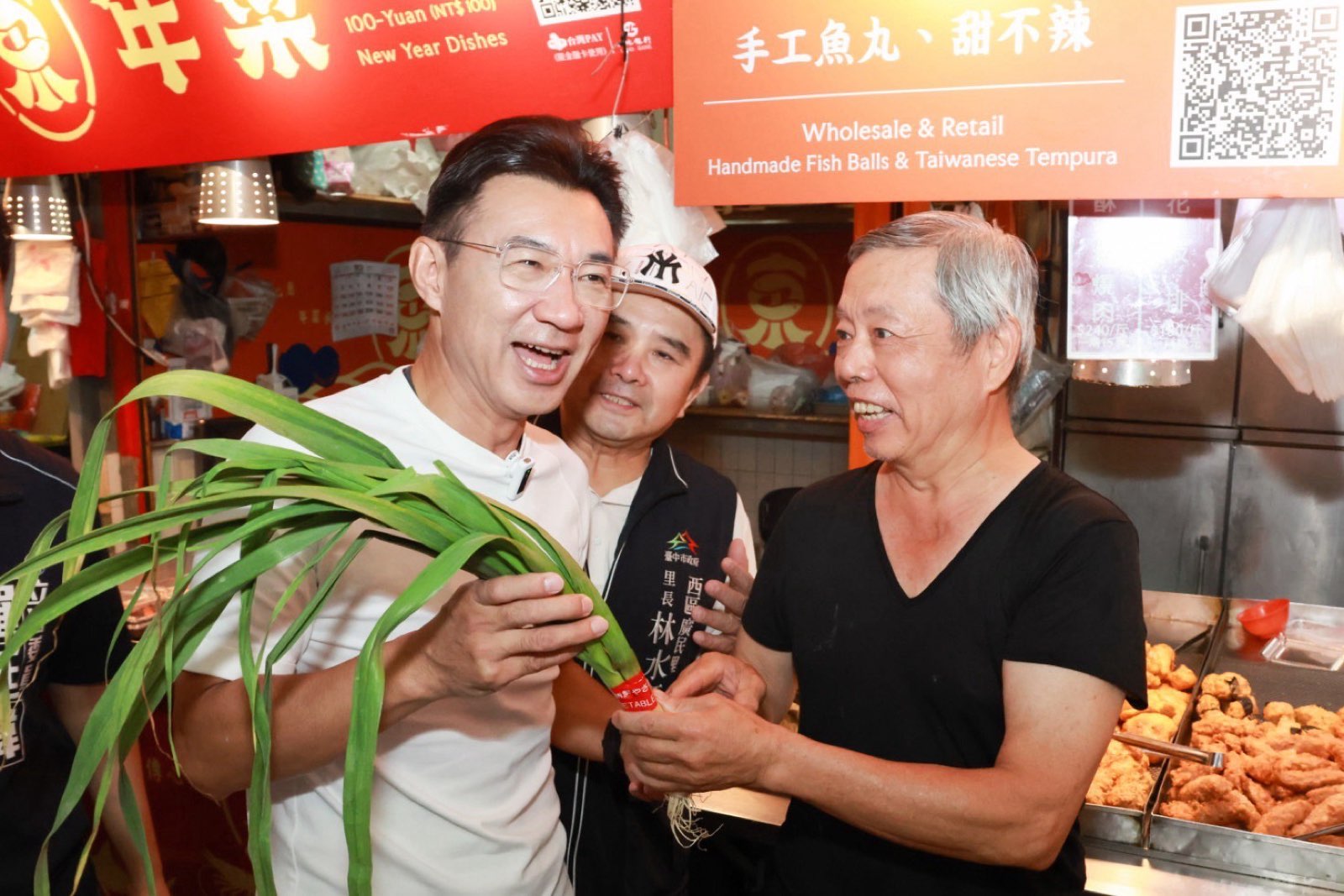 江啟臣在市場攤位手拿青蔥，與攤商及里長開心交談