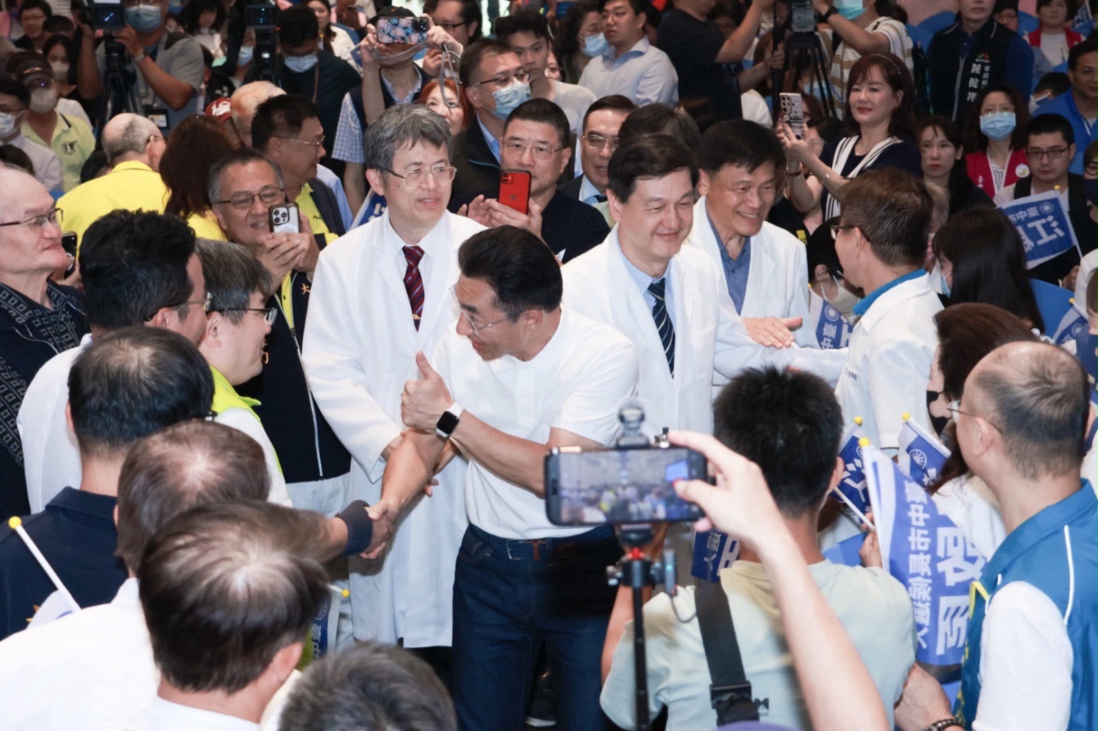 穿白色Polo衫的男子進場時與兩旁人士握手、比讚，周圍多位身穿白袍的醫事人員與民眾舉手機拍照，並揮舞印有「江啟臣」的藍旗