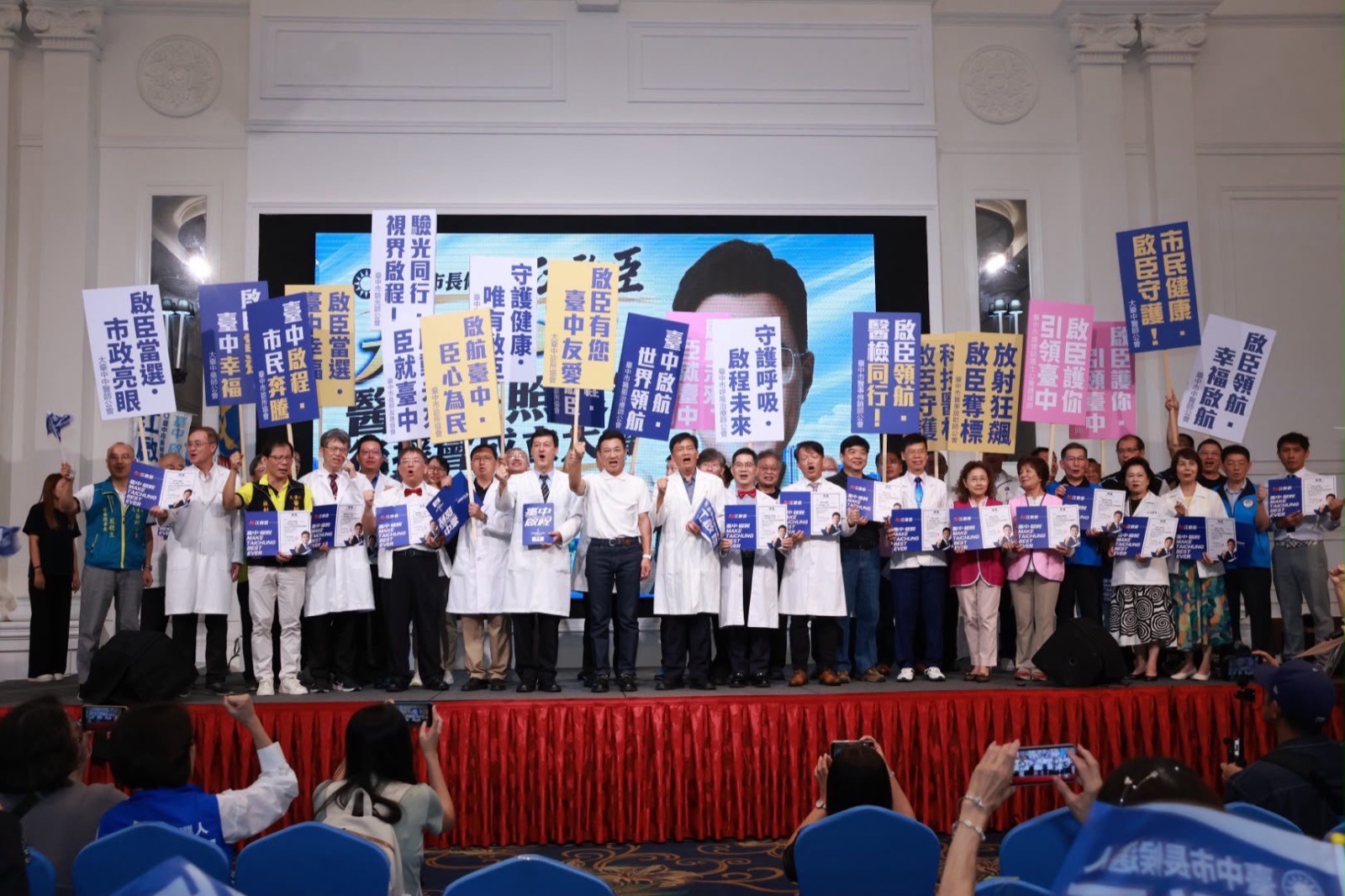 臺上眾人高舉「啟臣領航」「守護健康」「臺中啟程 市民奔騰」等各公會看板，前排穿白袍的醫事人員手持聘書，背後螢幕為江啟臣大幅照片