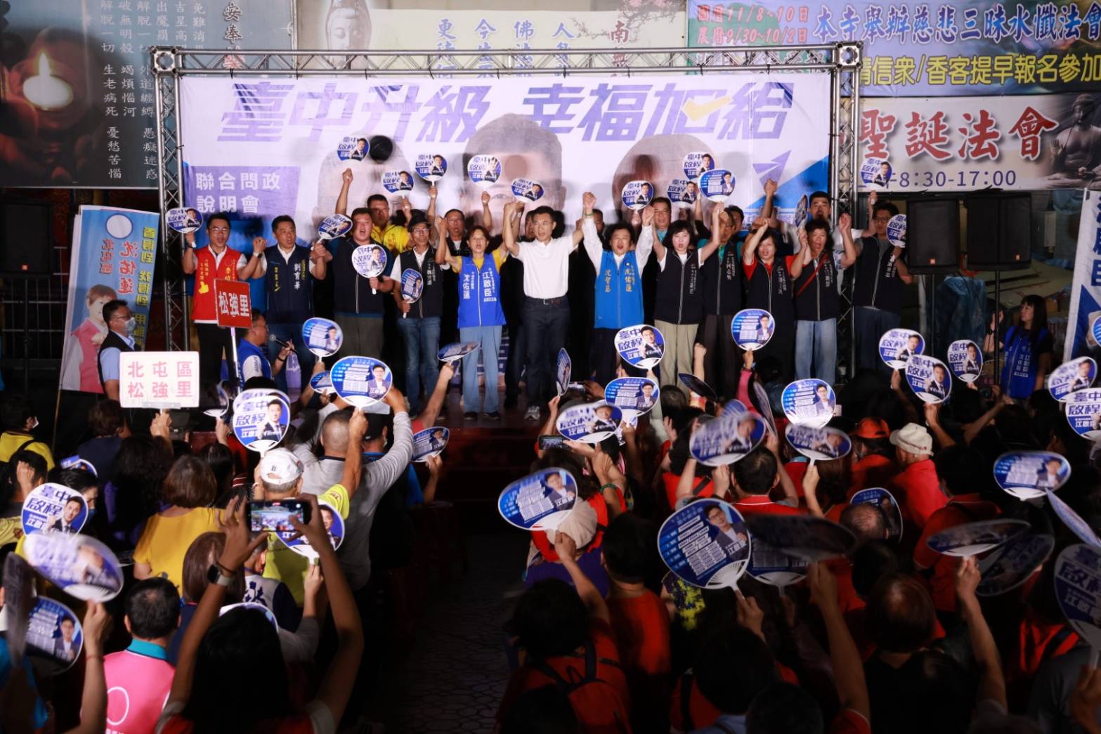 北屯聯合問政說明會臺上眾人手牽手高舉雙手，背心寫著「立法委員 黃健豪」「臺中市議員 沈佑蓮」，里長背心標示北屯區松茂里、松強里，臺下鄉親高舉競選扇