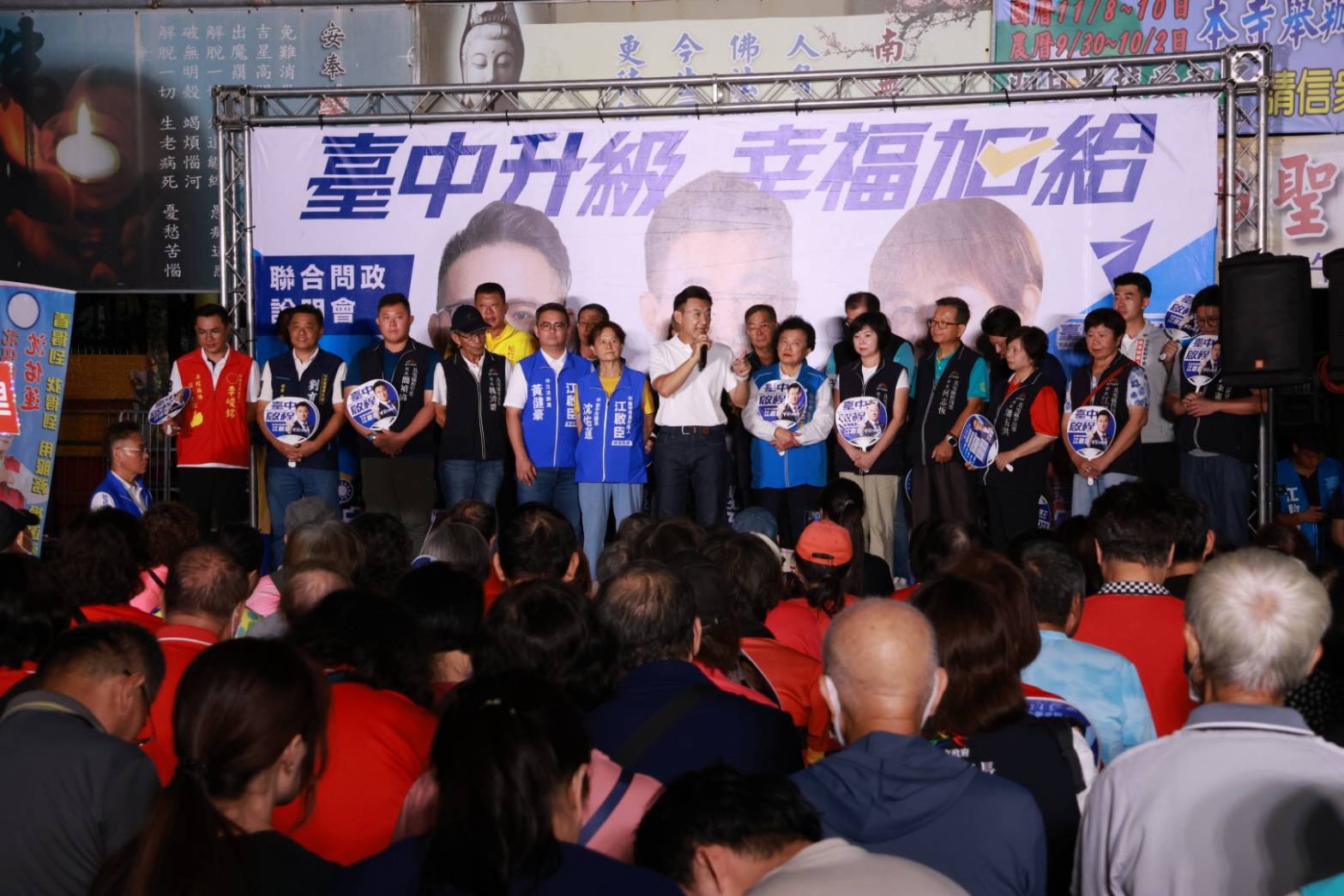 夜間廟埕前的聯合問政說明會，臺上嘉賓與里長排成一列，背板寫著「臺中升級 幸福加給」「聯合問政說明會」，臺下觀眾背對鏡頭聆聽