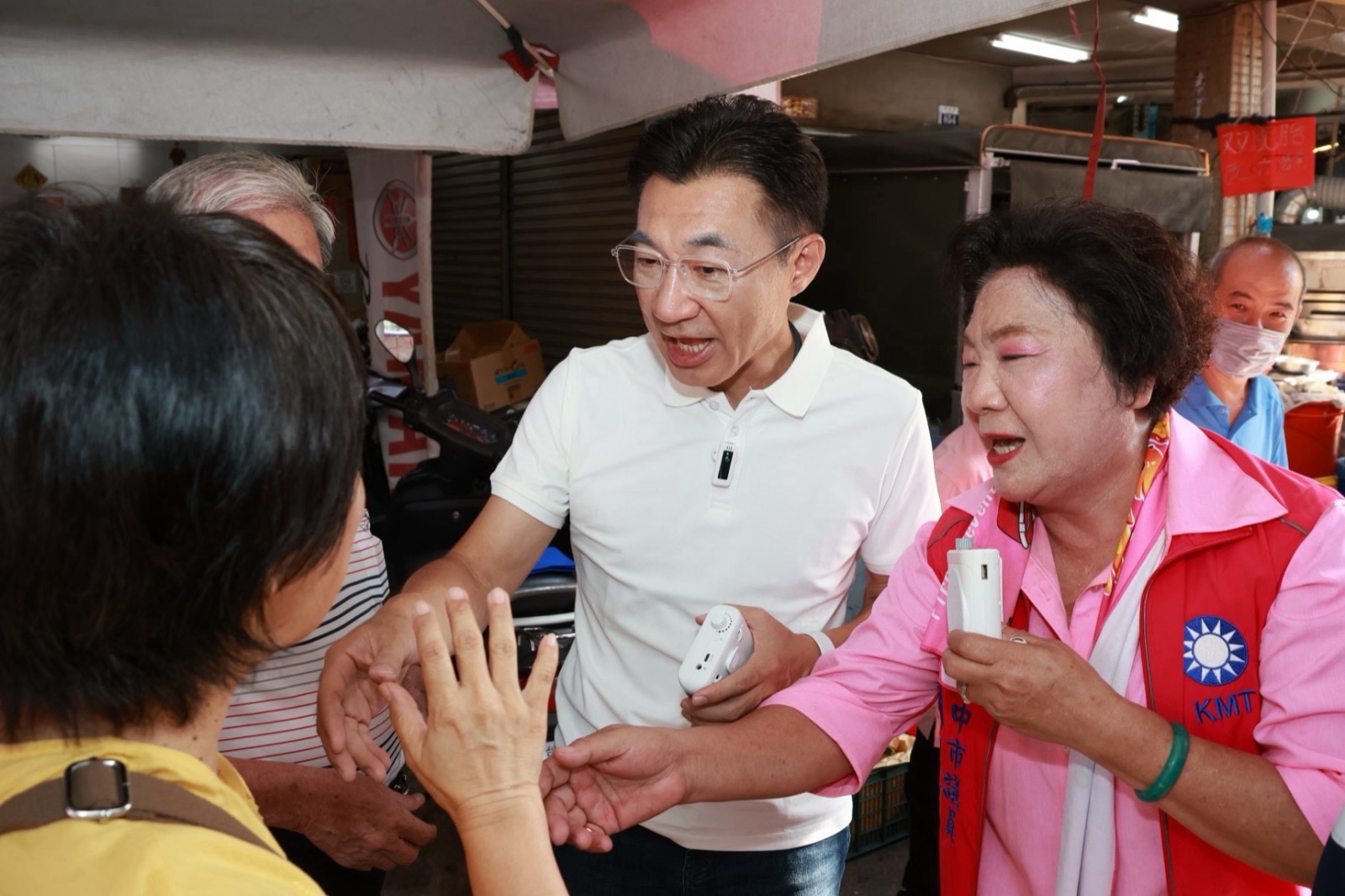 穿白色Polo衫的男子在市場攤位間與民眾握手交談，右側穿粉紅襯衫、紅色國民黨背心的女士也向民眾伸手致意