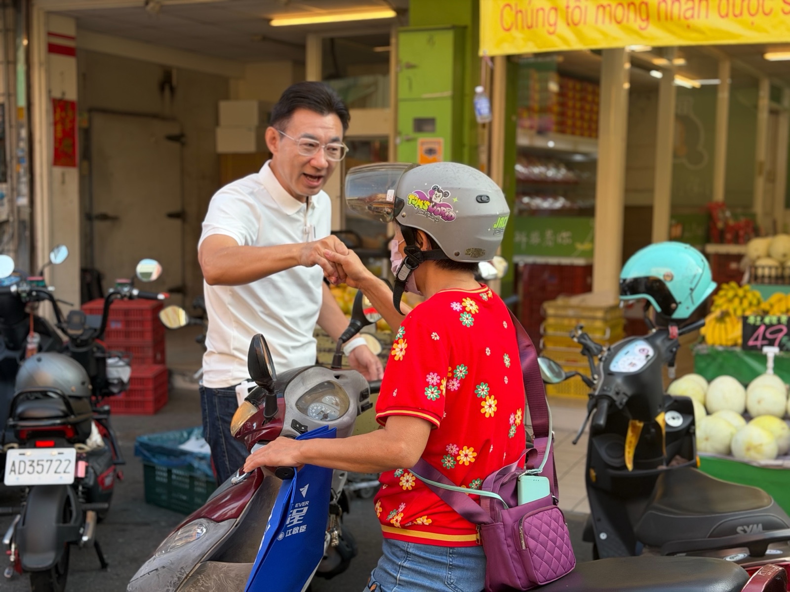 穿白色Polo衫的男子在水果攤前與一位戴安全帽、坐在機車上的女士碰拳，機車上放著「臺中啟程 江啟臣」藍色提袋