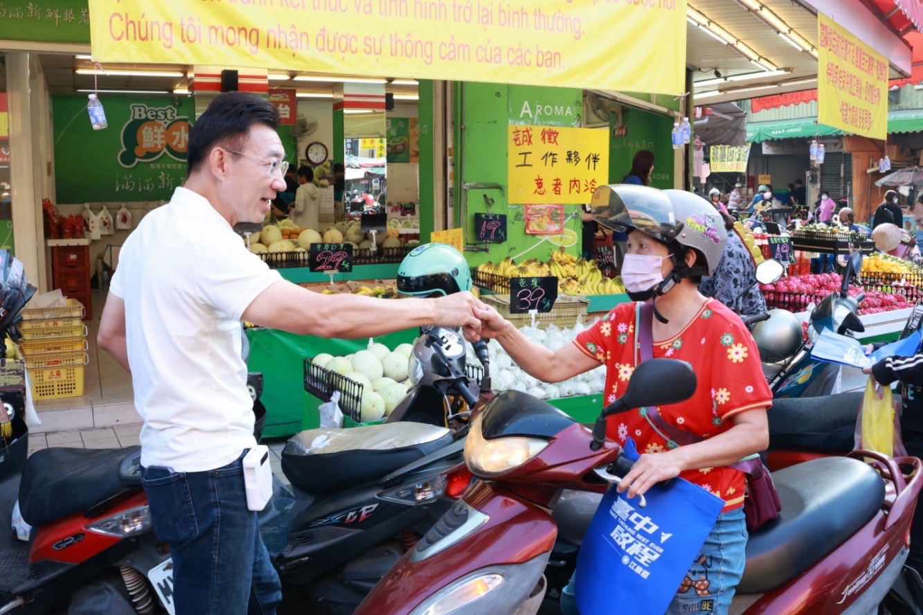 穿白色Polo衫的男子在市場路邊與一位戴安全帽、騎著機車的女士握手，女士車前掛著「臺中啟程 江啟臣」藍色提袋，後方是水果攤
