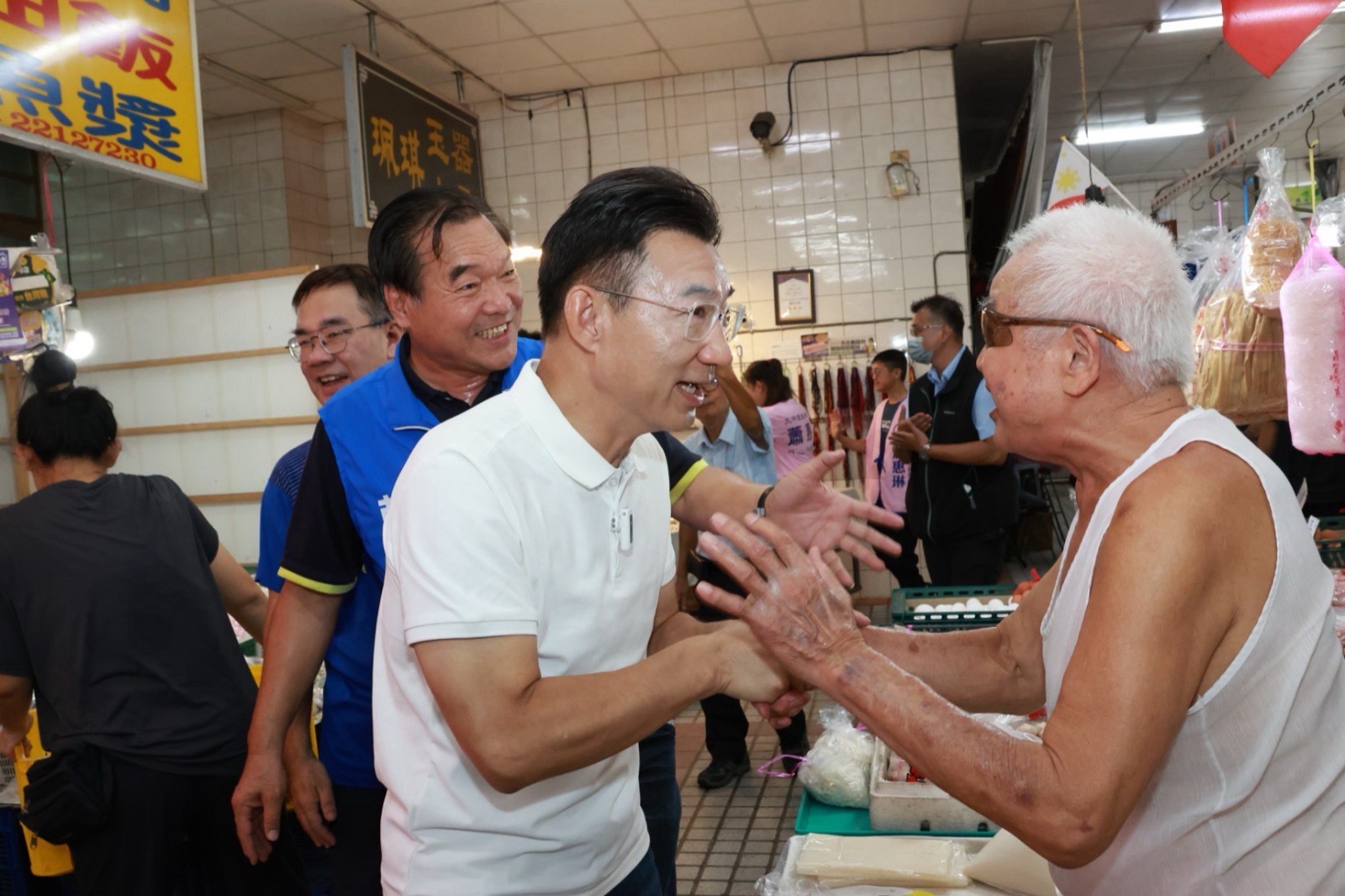 穿白色Polo衫的男子在傳統市場內雙手握住一位穿白色背心的攤商長者的手，身後穿藍色背心的男子笑著陪同