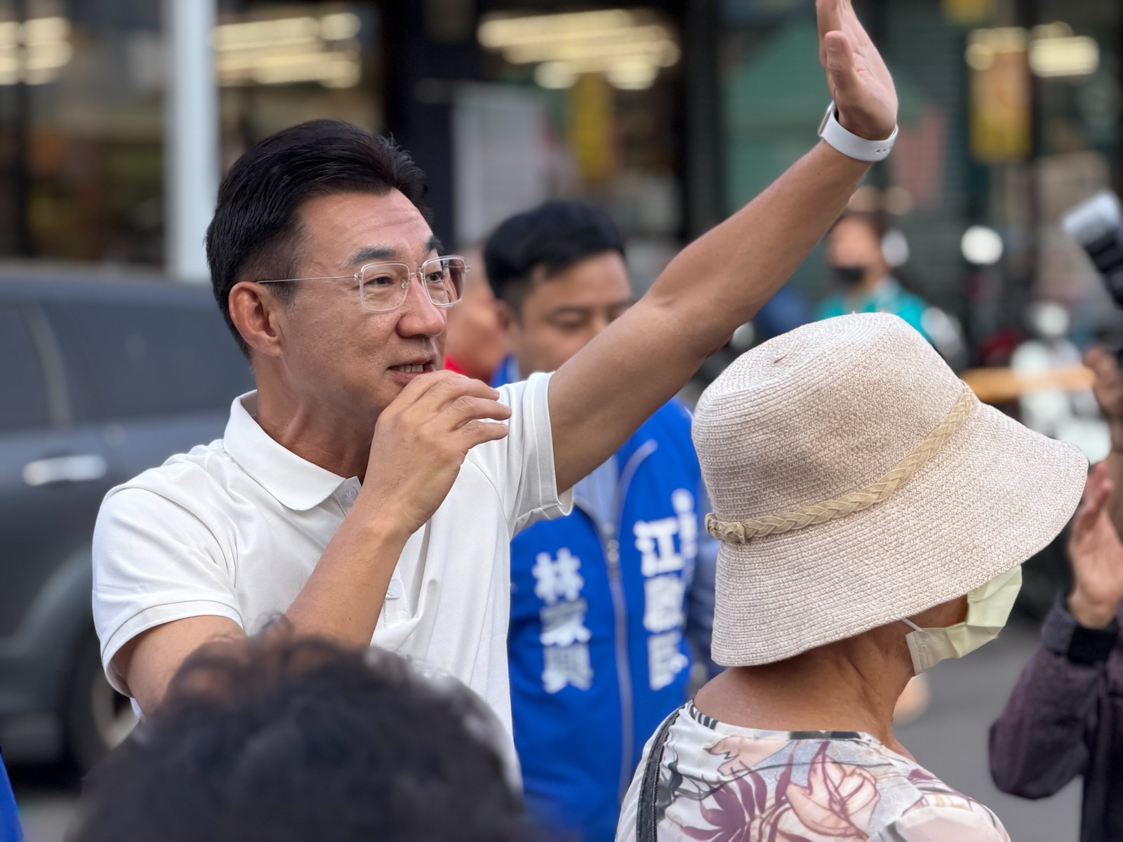 穿白色Polo衫的男子在街頭高舉右手向群眾揮手，身後穿藍色「江啟臣」背心的人在側，前方戴遮陽帽的長輩背對鏡頭