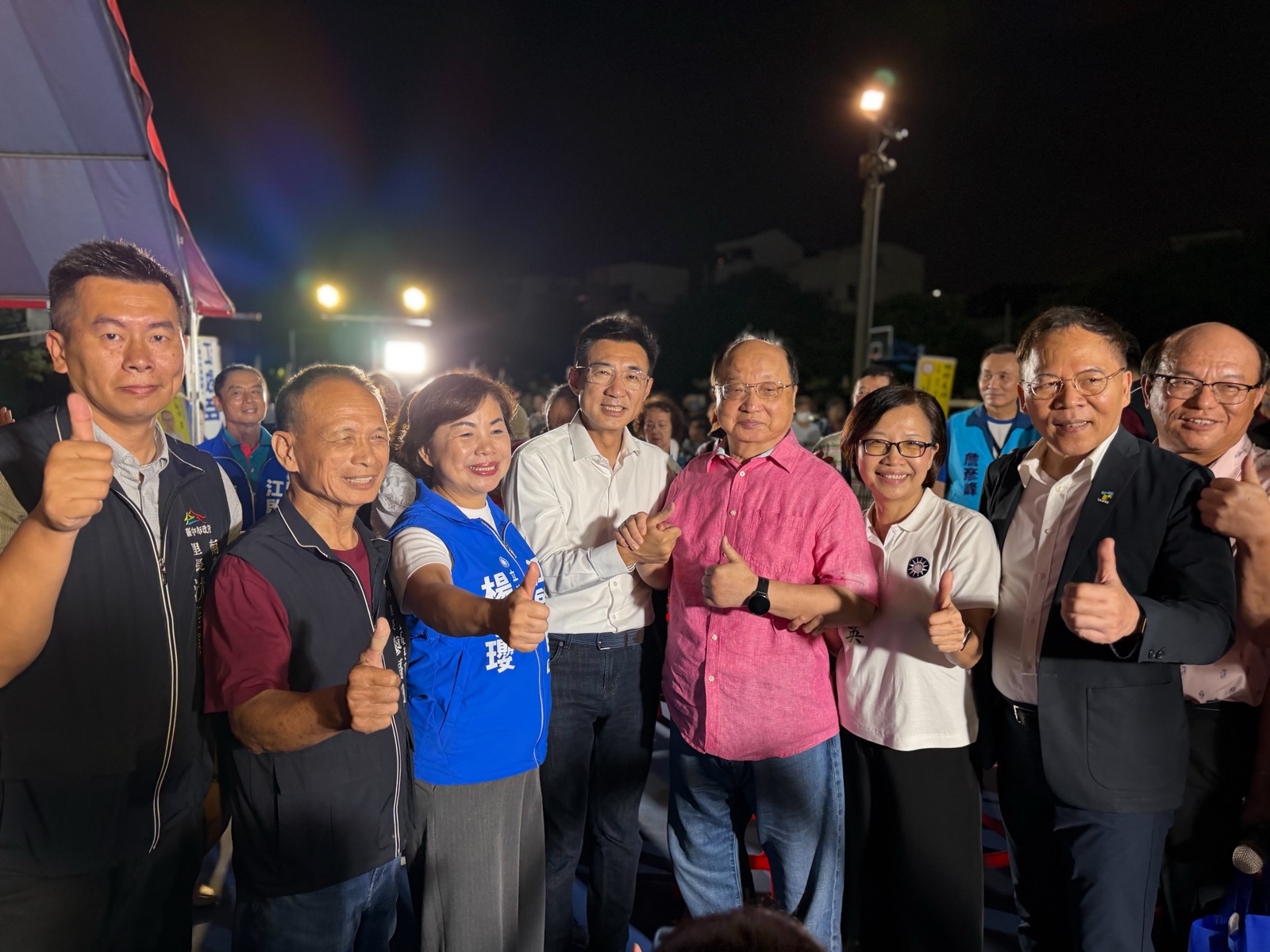 夜間球場邊，穿白襯衫的男子與穿粉紅襯衫的長者、穿藍色「立法委員」背心的女士、穿白色Polo衫的女士及多位民眾合影比讚