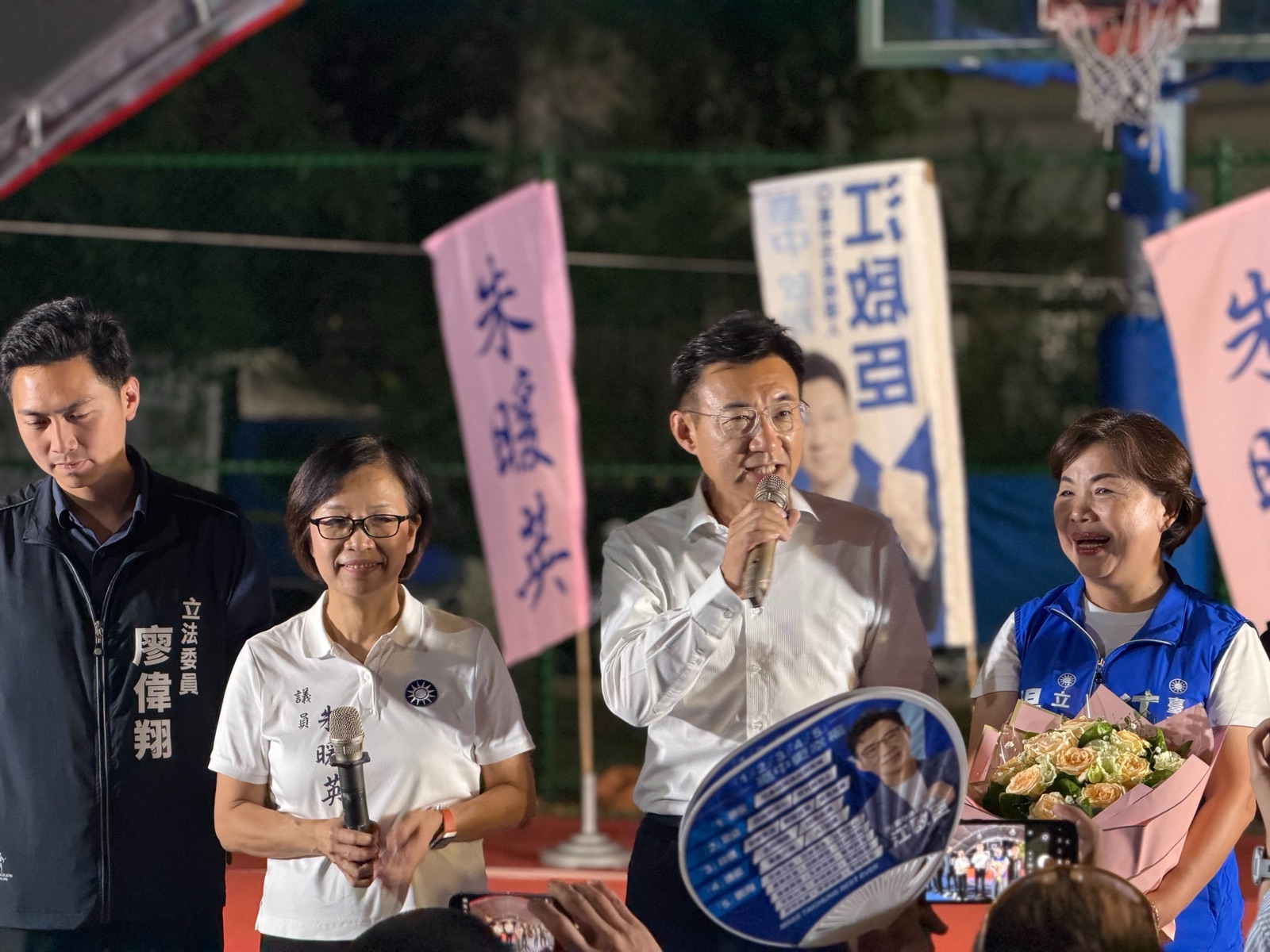 穿白襯衫的男子手持麥克風發言，左側背心寫著「立法委員 廖偉翔」，身旁穿白色Polo衫的女士衣上繡有「議員 朱暖英」，右側穿藍色背心的女士手捧花束