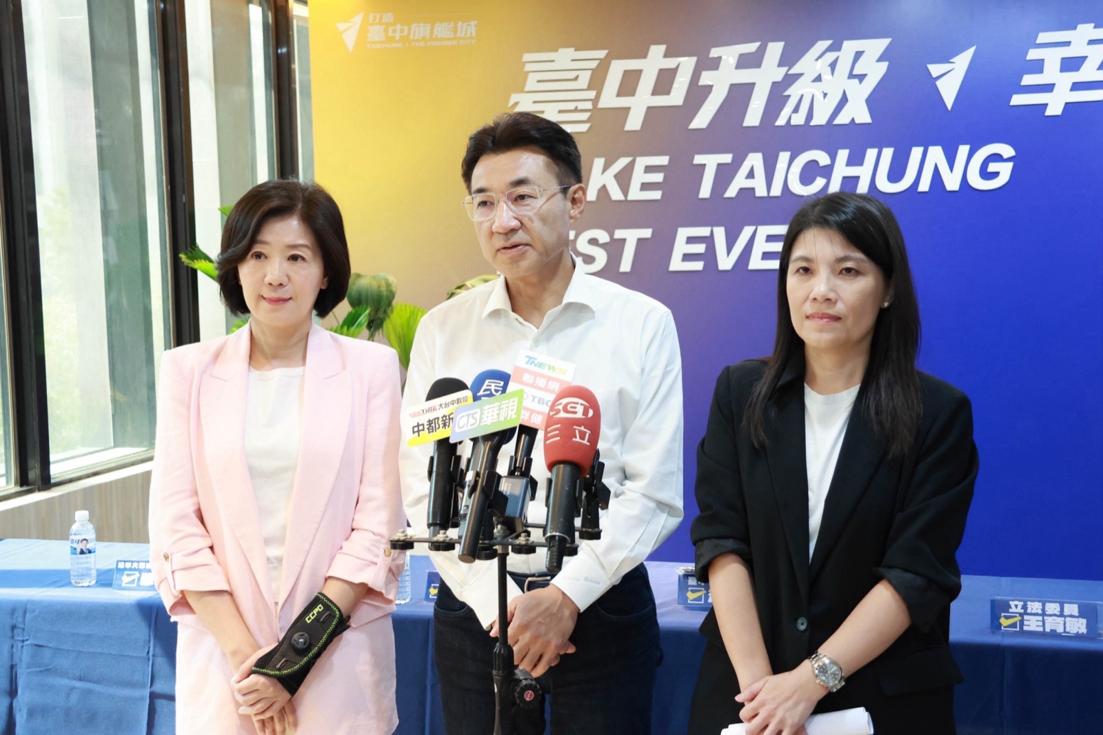 記者會後，穿白襯衫的男子面對多家媒體麥克風受訪，兩旁各站一位女士，背景為「臺中升級 幸福加給」背板