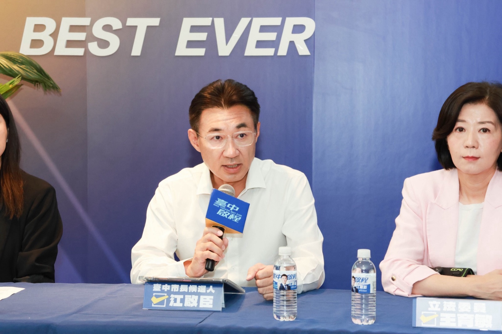 穿白襯衫的男子手持麥克風發言，桌牌寫著「臺中市長候選人 江啟臣」，右側桌牌寫著「立法委員 王育敏」