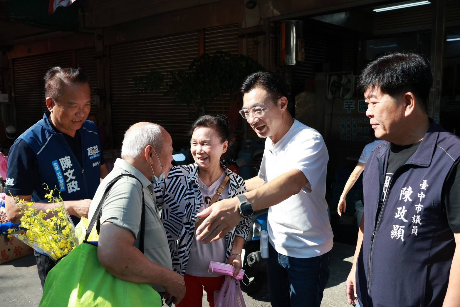 市場街口，穿白色上衣的男子伸手向採買的民眾問好，左側穿藍色「陳文政」背心、右側穿「臺中市議員 陳政顯」背心的男子在旁陪同