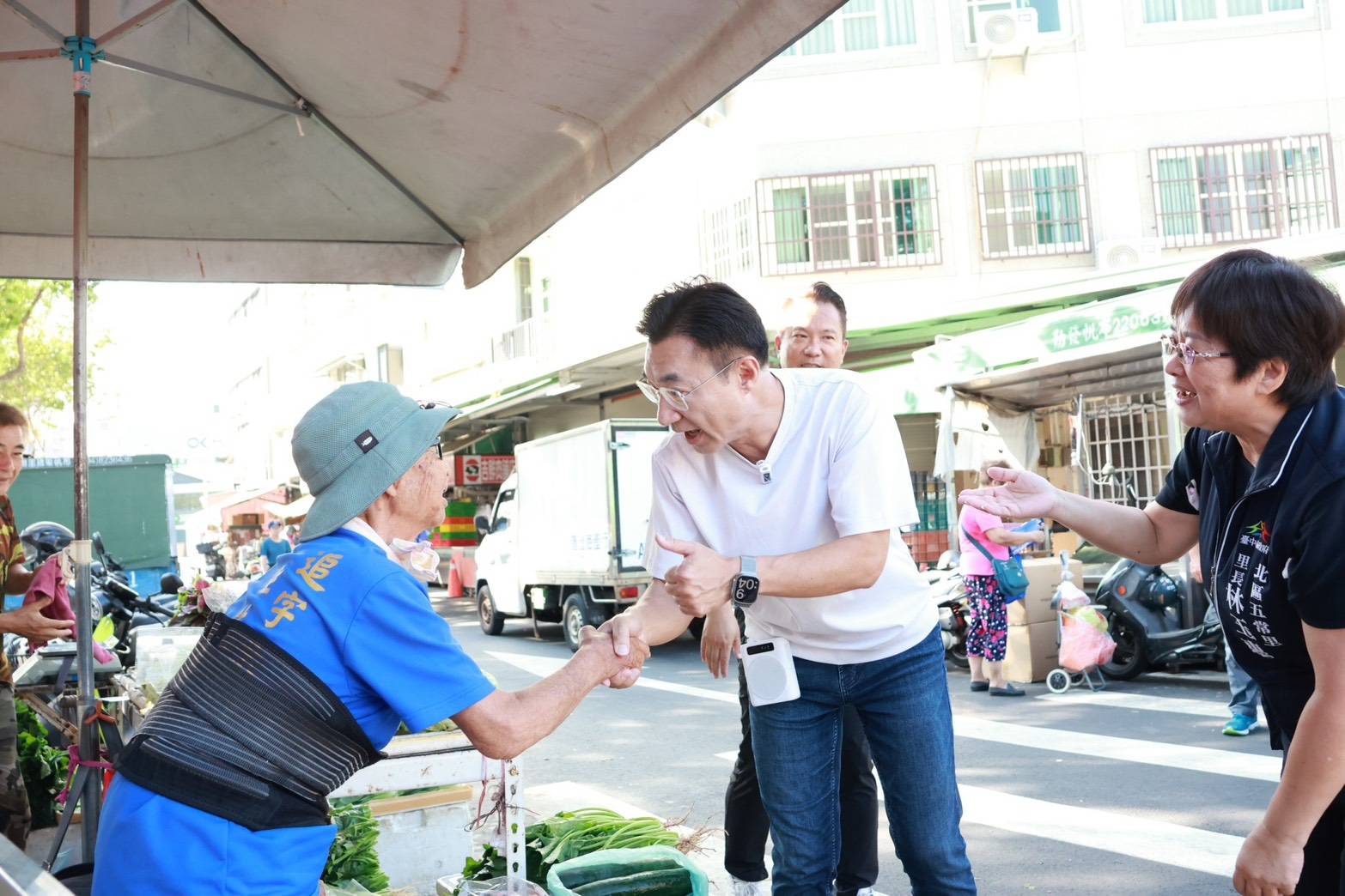 穿白色上衣的男子在菜攤前與一位戴帽的長輩握手，右側穿「北區五常里 里長」背心的女士伸手致意