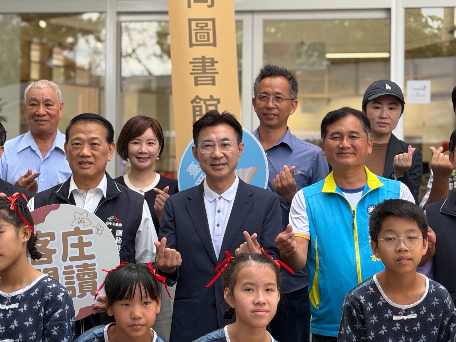 開館典禮合影特寫，穿深色西裝外套的男子與身旁嘉賓比愛心手勢，前方穿客家藍染花布衫的學童，後方可見「客庄閱讀」立牌與「石岡圖書館」布條